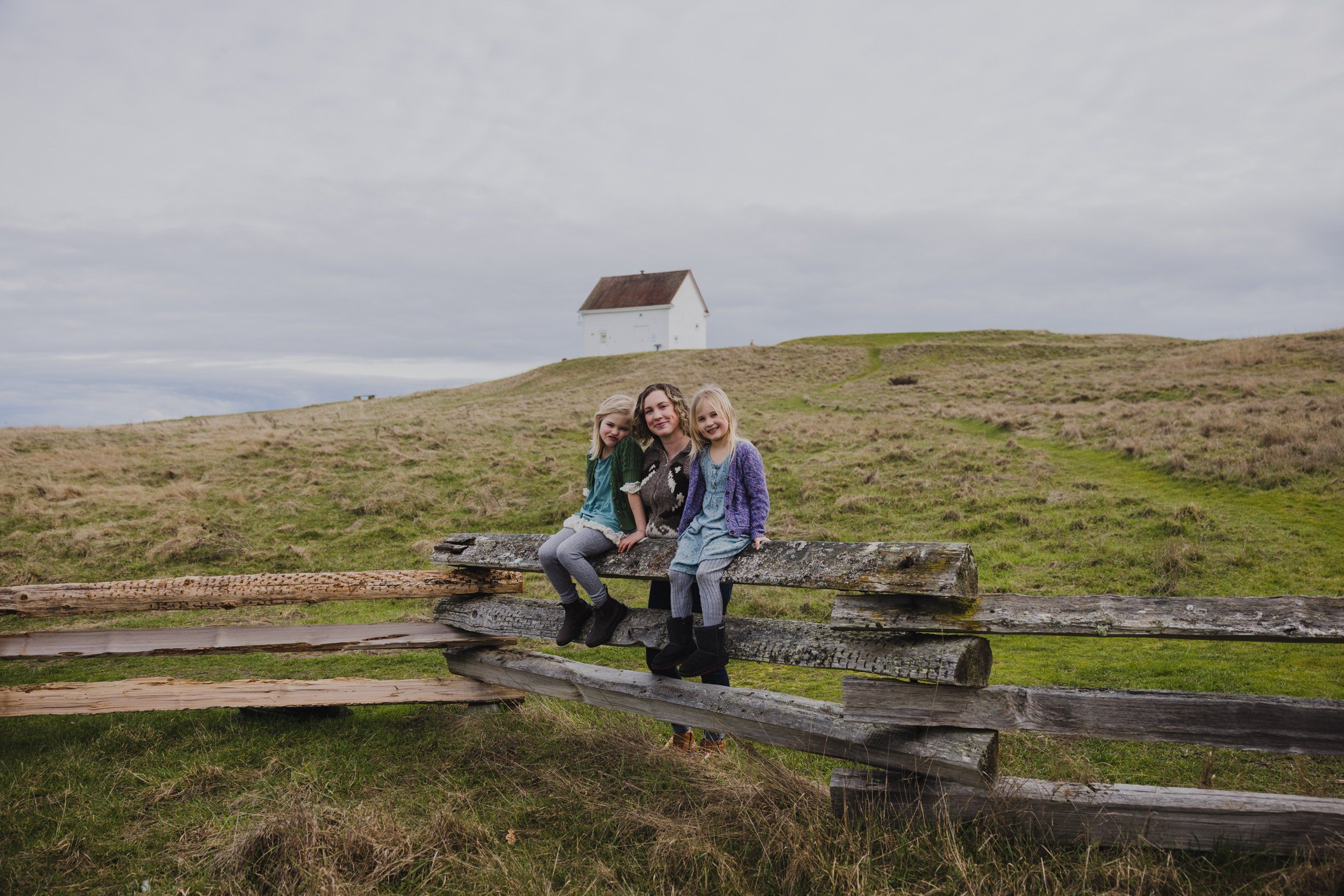 Three people sit on weathered wooden fence rails in a green meadow with a distant cabin and overcast sky