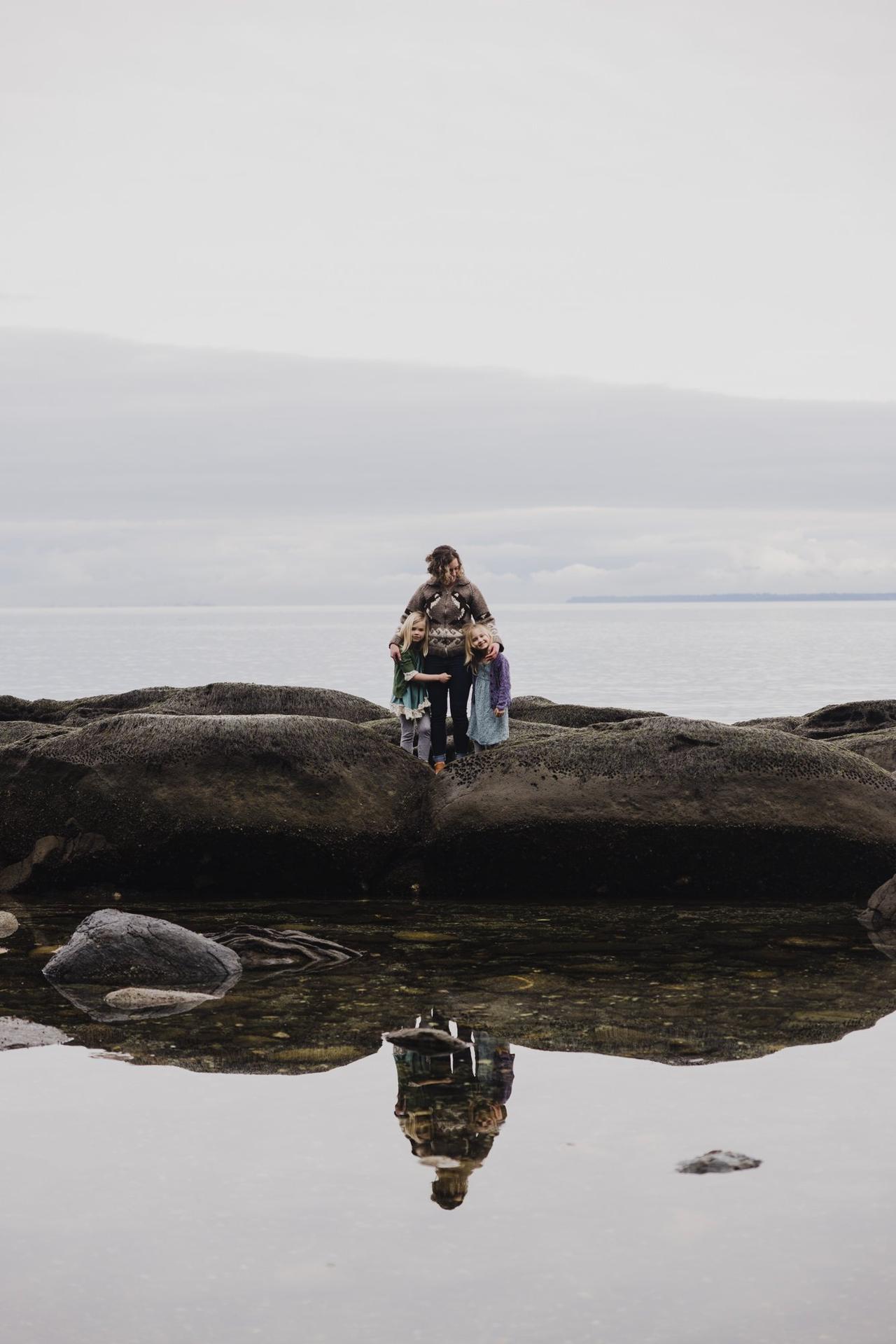 Adult and two children stand on moss-covered rocks above a tidal pool reflecting their figures on Saturna Island