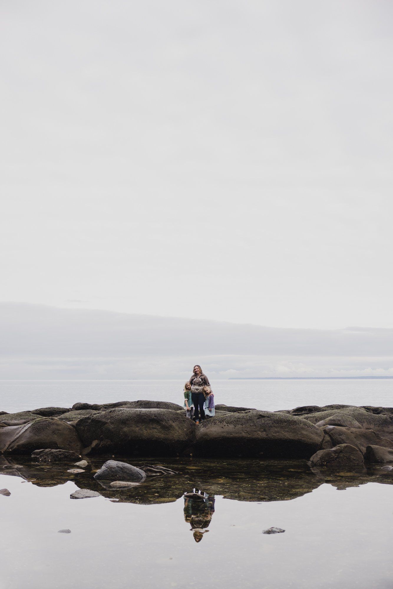 Woman seated on rocky shoreline reflected in still tide pool, misty water and hills beyond