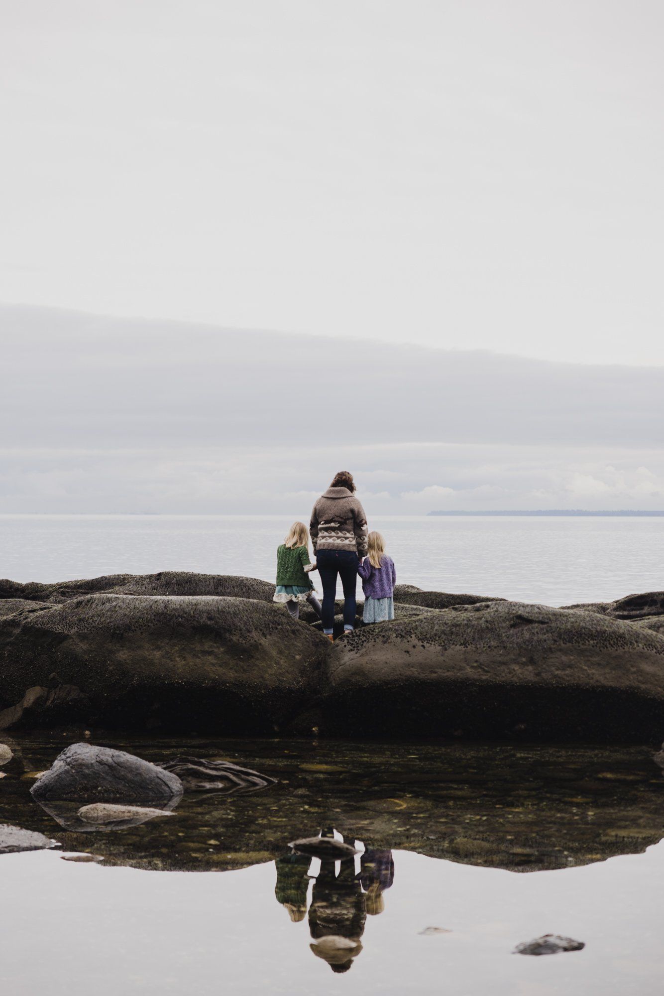 Family of three standing on a driftwood log reflected in still water, overcast coastal landscape behind them
