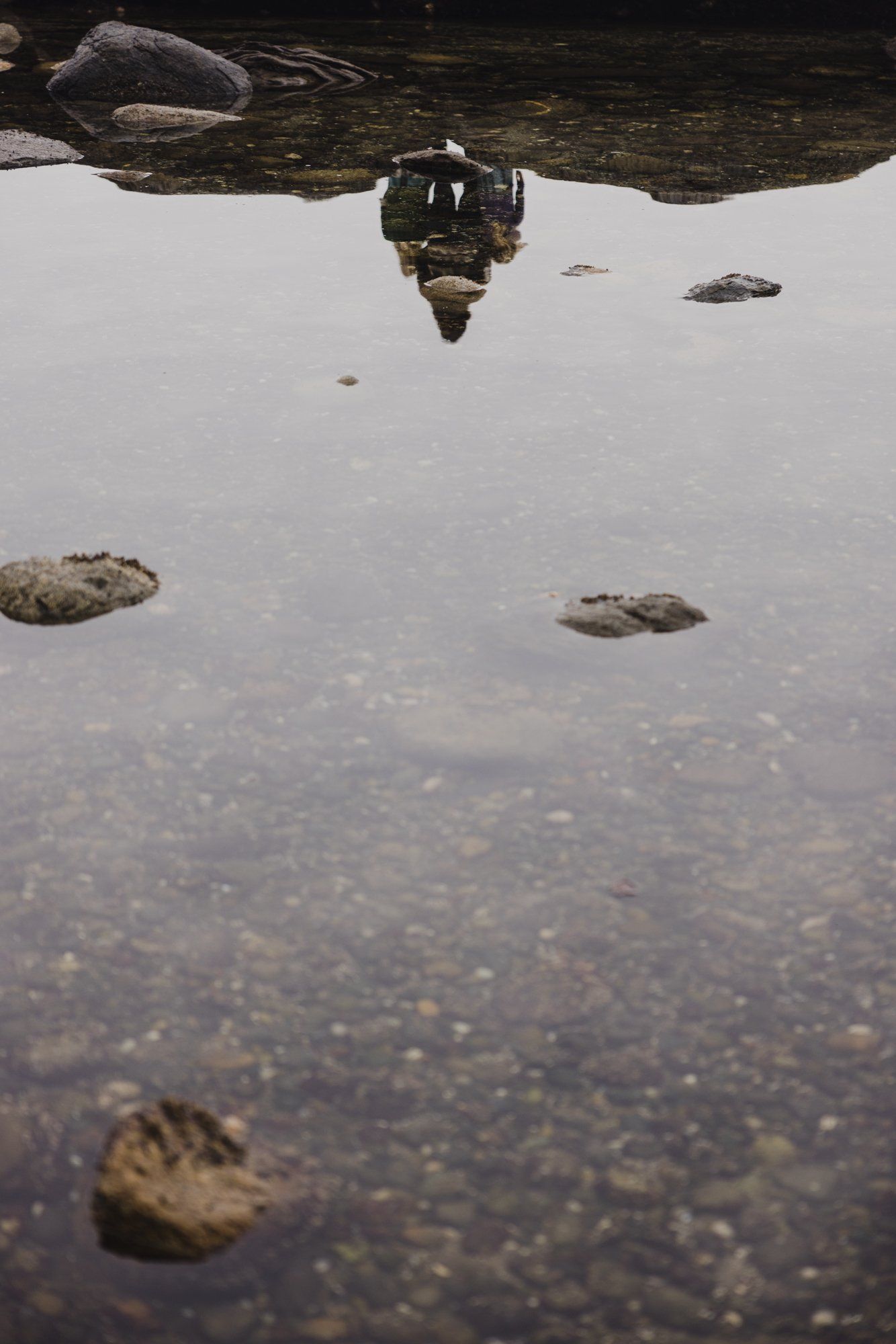 Reflection of a person in shallow tidal water surrounded by smooth stones on a rocky shore