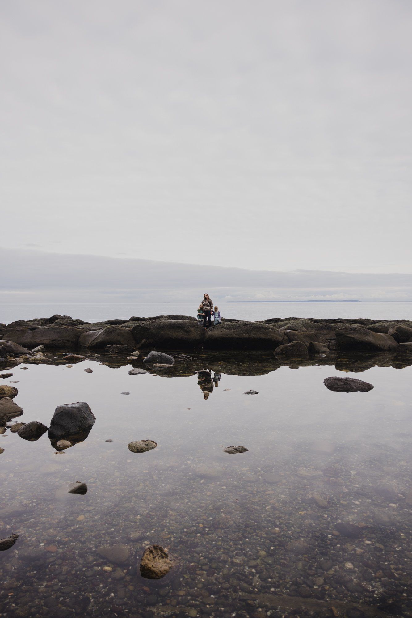 Woman seated on rocky outcrop reflected in still tidal pool beneath overcast sky