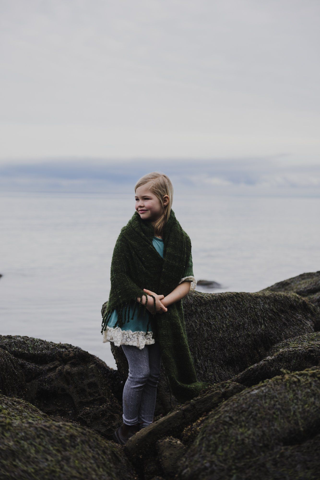 Young girl in dark cardigan and blue dress standing on rocky shoreline with calm ocean and overcast sky behind her