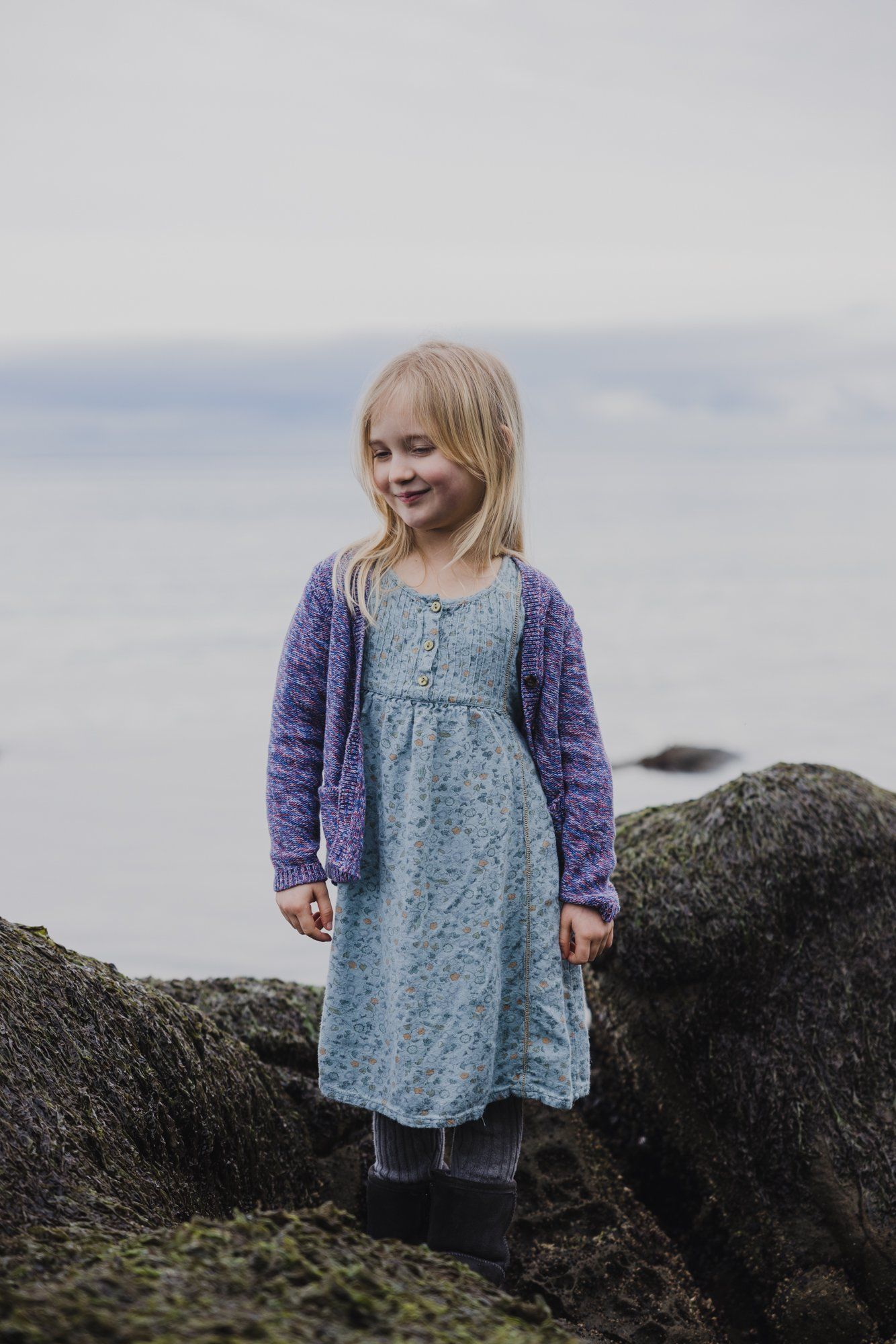 Young girl with blonde hair standing on moss-covered rocks, wearing a blue dress and purple cardigan, gazing to the side with ocean horizon behind her