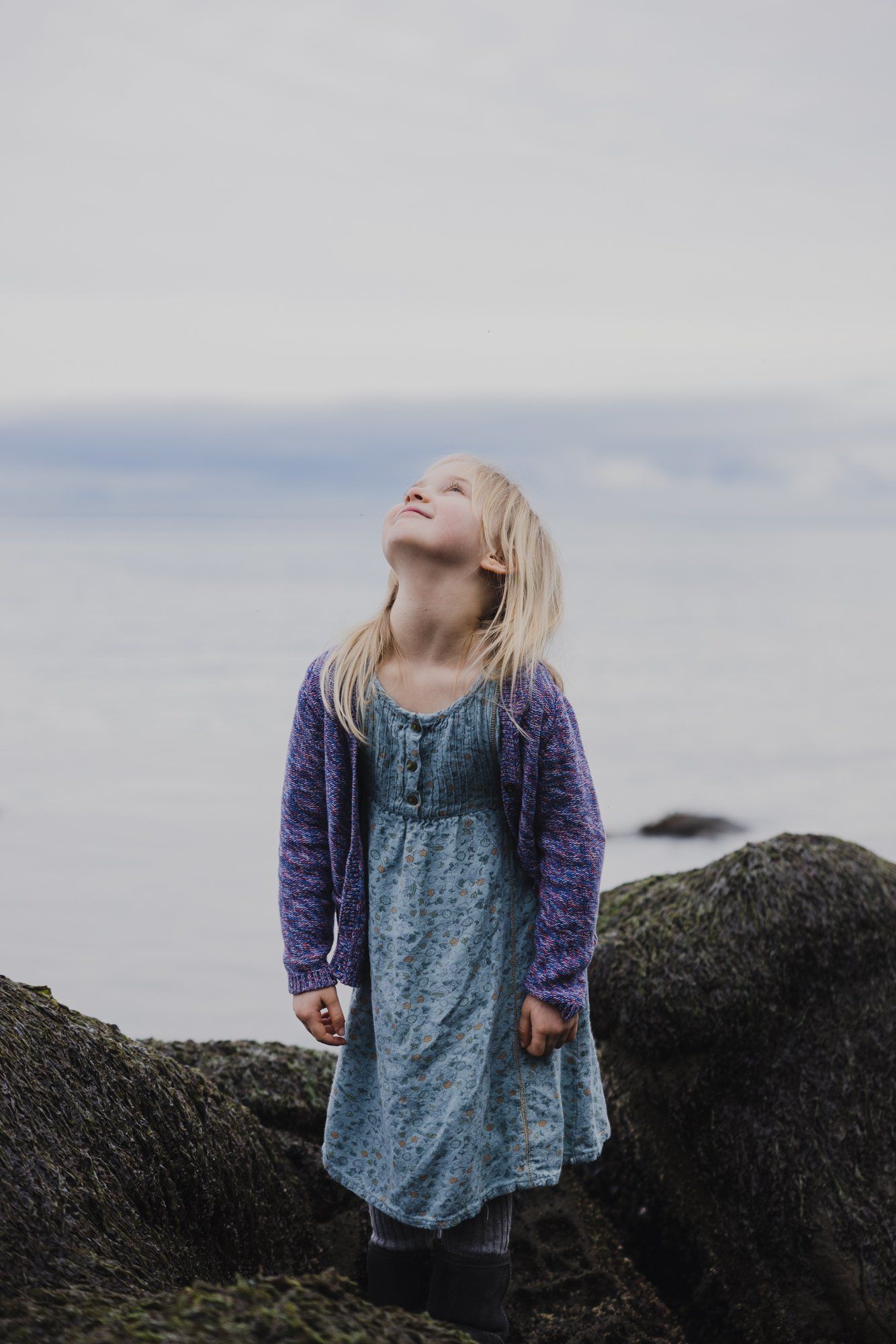 Young girl with blonde hair looking upward, standing on dark rocks with overcast sky and calm water behind her
