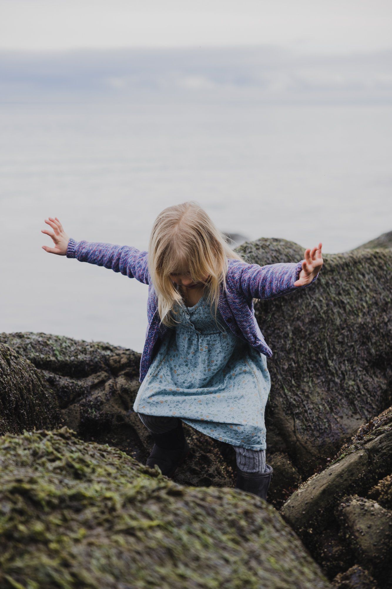 Young child with arms spread wide standing among moss-covered rocks under overcast coastal sky