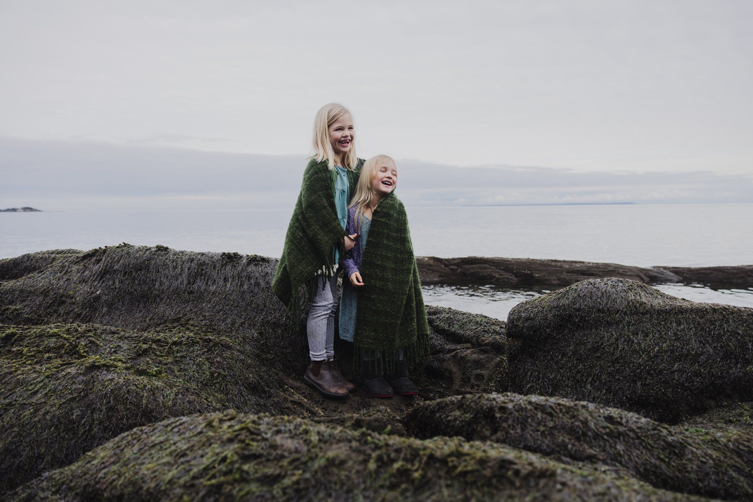 Woman and child wrapped in moss-green blankets stand on rocky, lichen-covered shores with misty water beyond