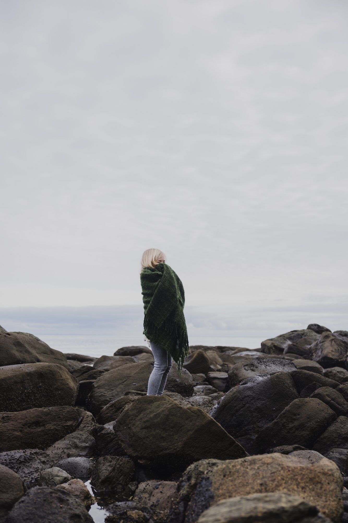 Woman in dark jacket standing on rocky shoreline with calm ocean and overcast sky behind her