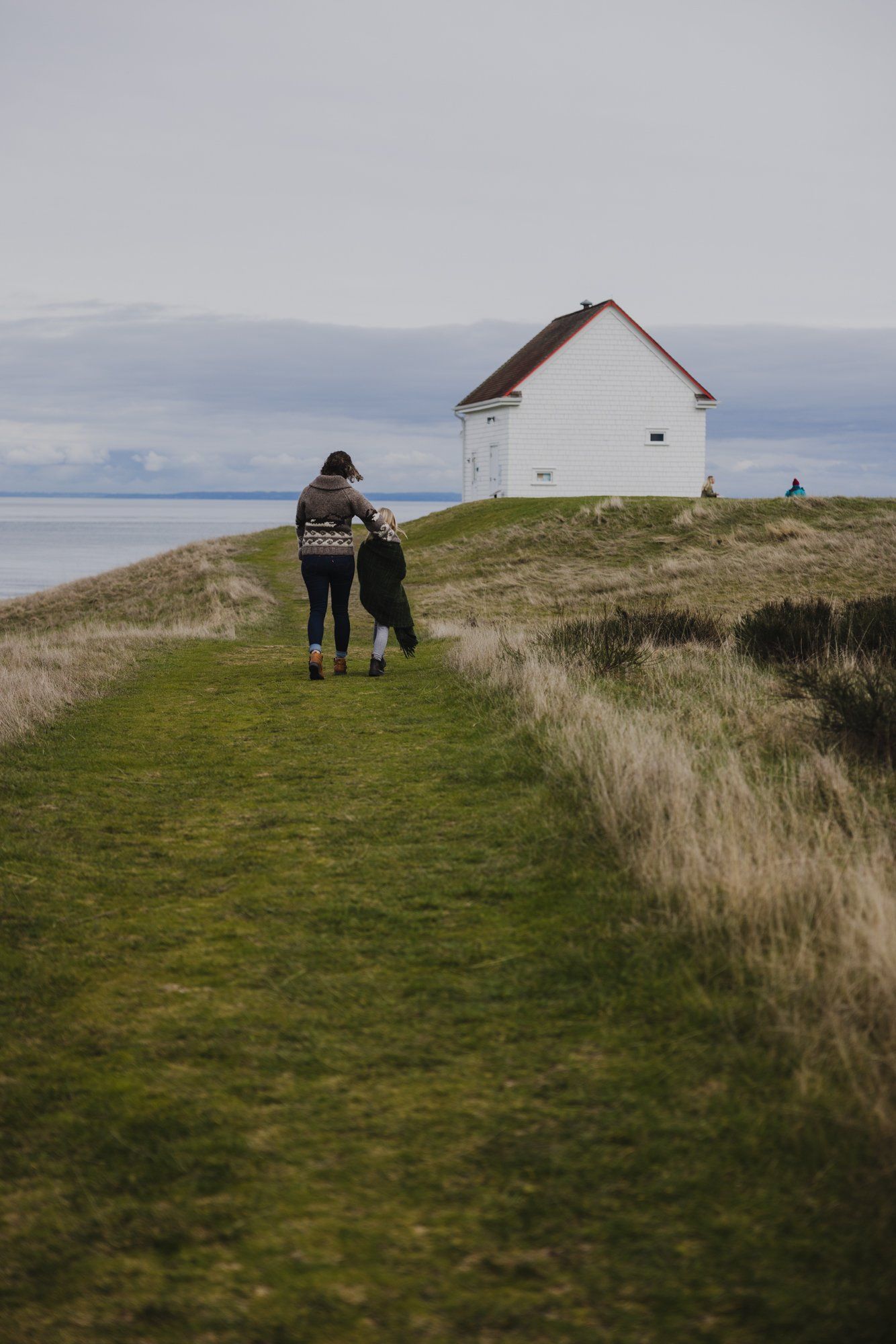 Two people walk toward a white house with a red roof on a grassy bluff overlooking calm water under overcast skies