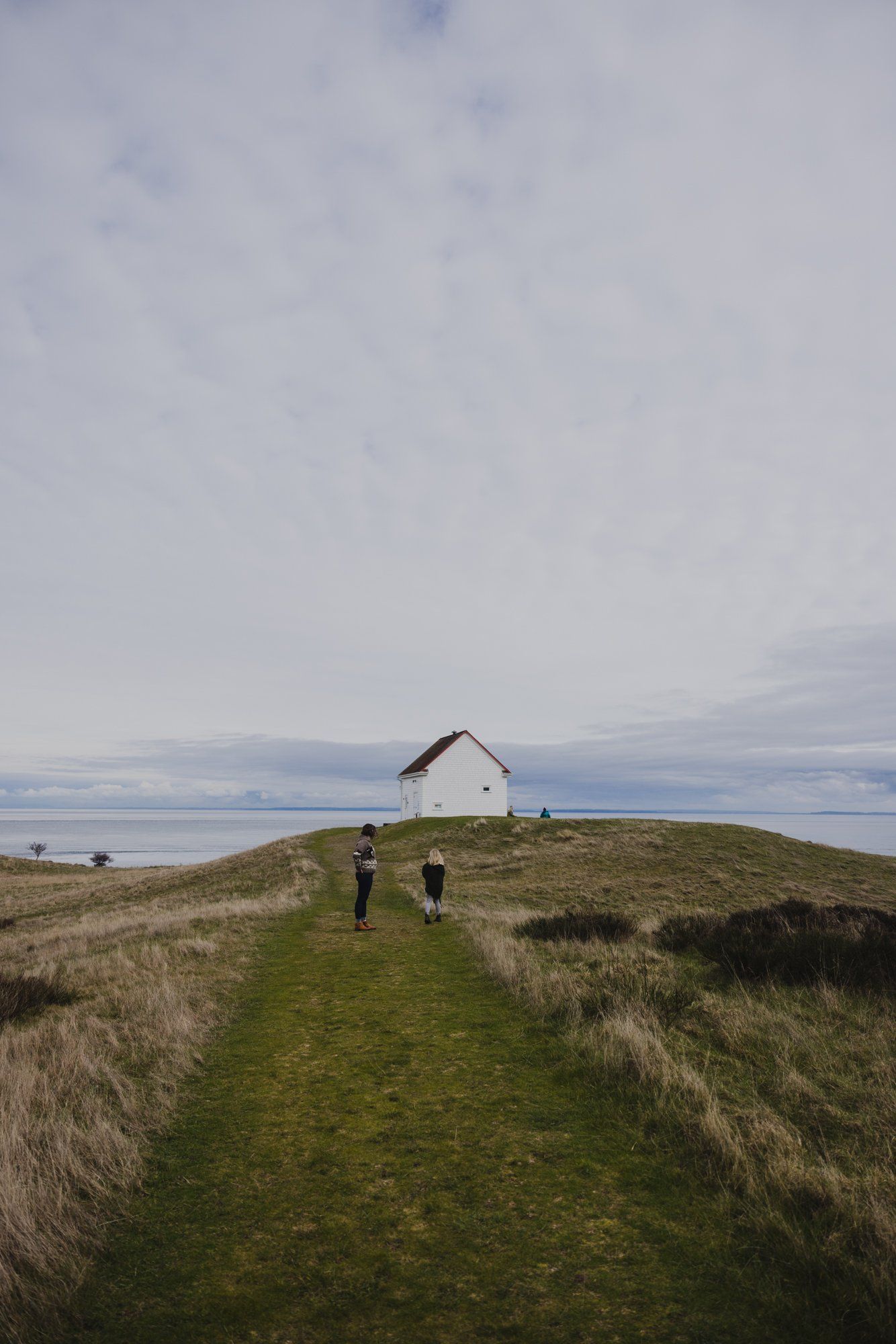 Two figures walk toward a solitary white house on a grass-covered hilltop under moody, overcast skies