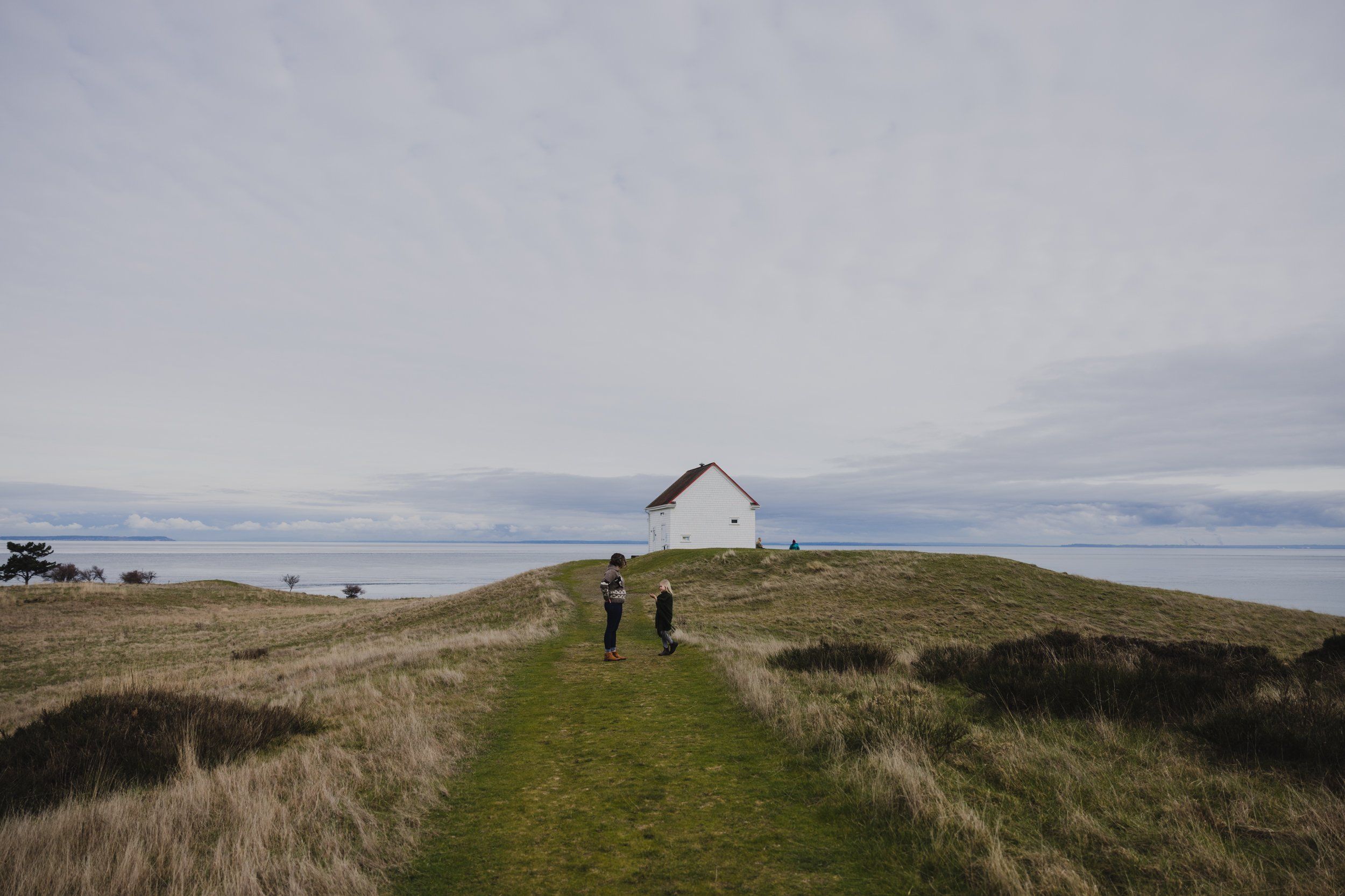 Family walking toward a white house on a grassy hilltop overlooking the ocean under overcast skies