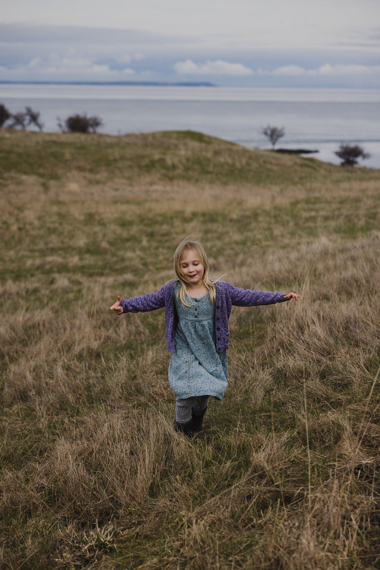 Young girl with arms spread wide in a grassy field overlooking calm water under cloudy skies