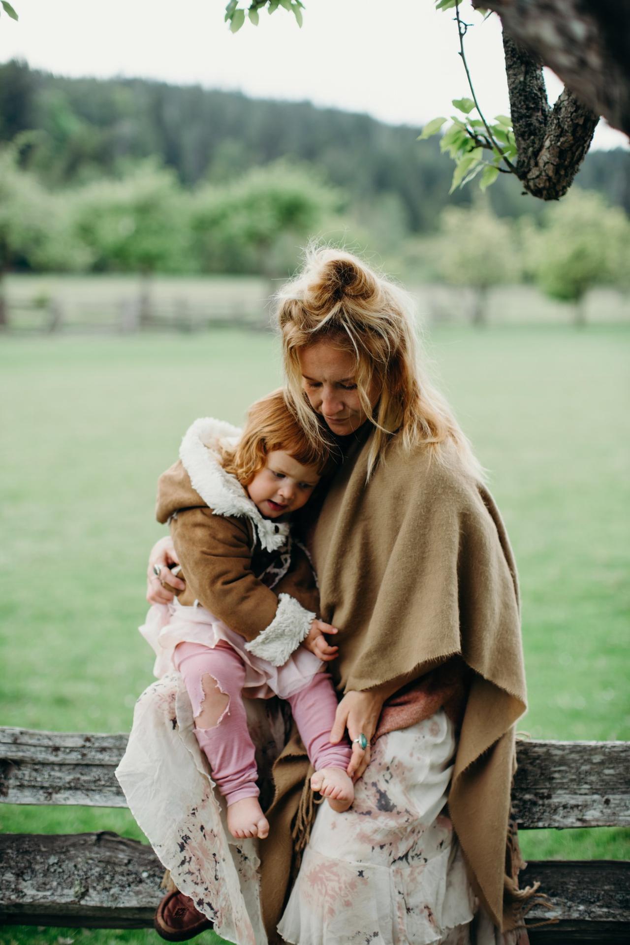 Woman and toddler embrace on a rocky shore with forested hills and water blurred behind them on Salt Spring Island