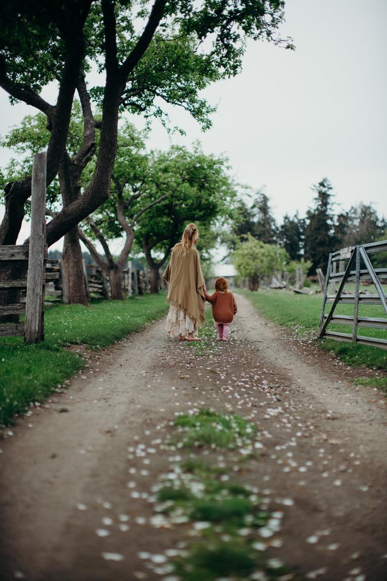 Woman and toddler holding hands on a tree-lined gravel path, soft overcast light filtering through green canopy on Salt Spring Island Farm