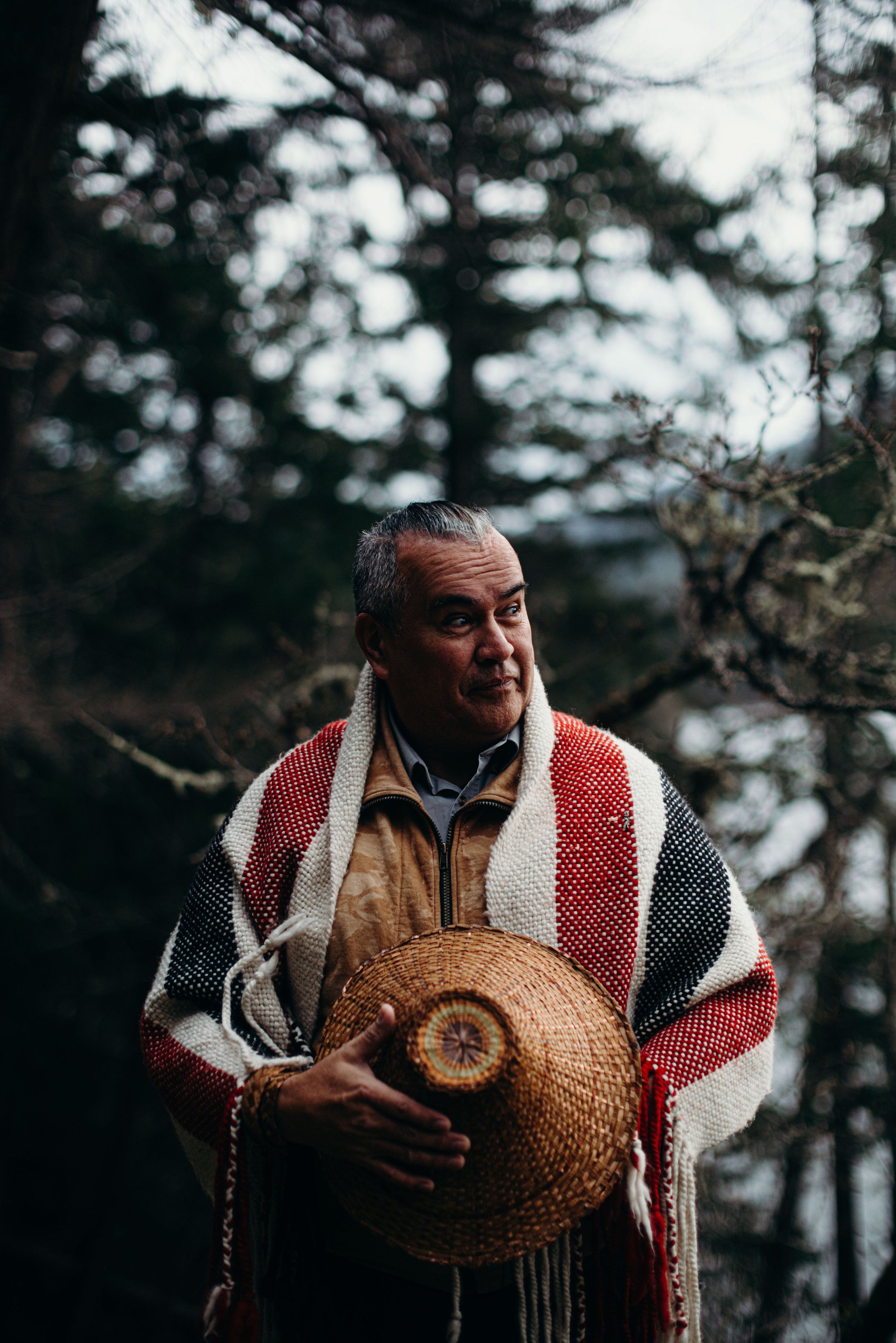 Man in red and white striped blanket holds woven basket against forested backdrop in natural daylight