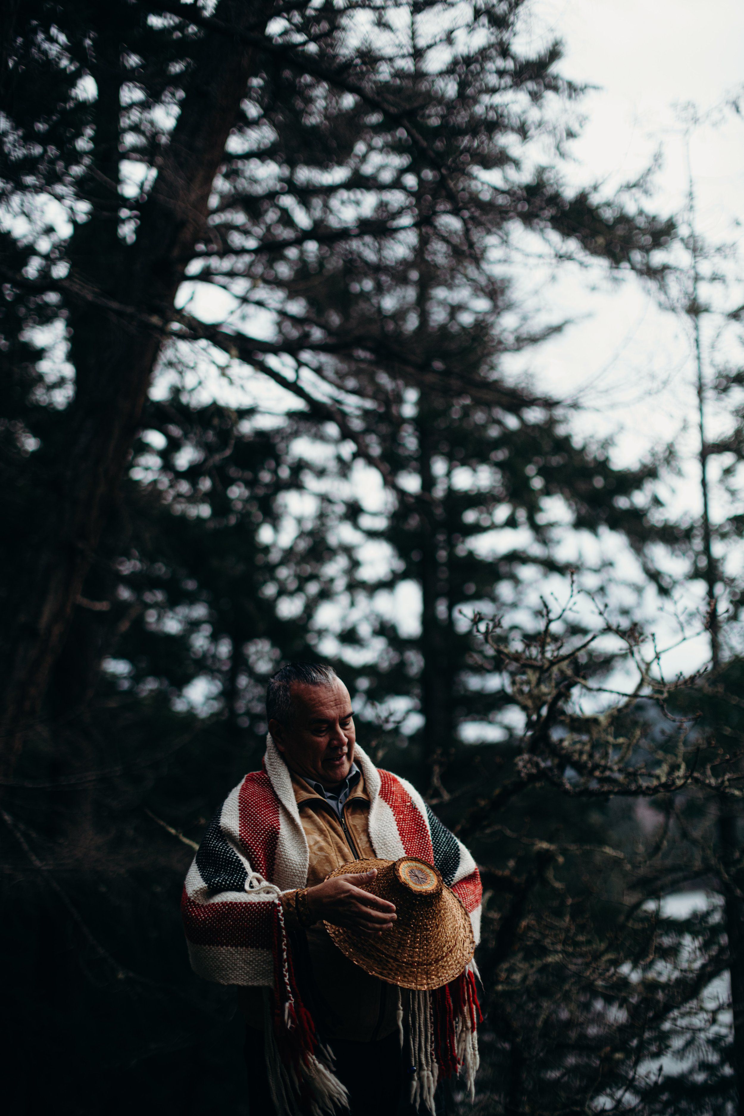 Indigenous man in red shirt holding woven basket stands among tall evergreen trees in dappled forest light