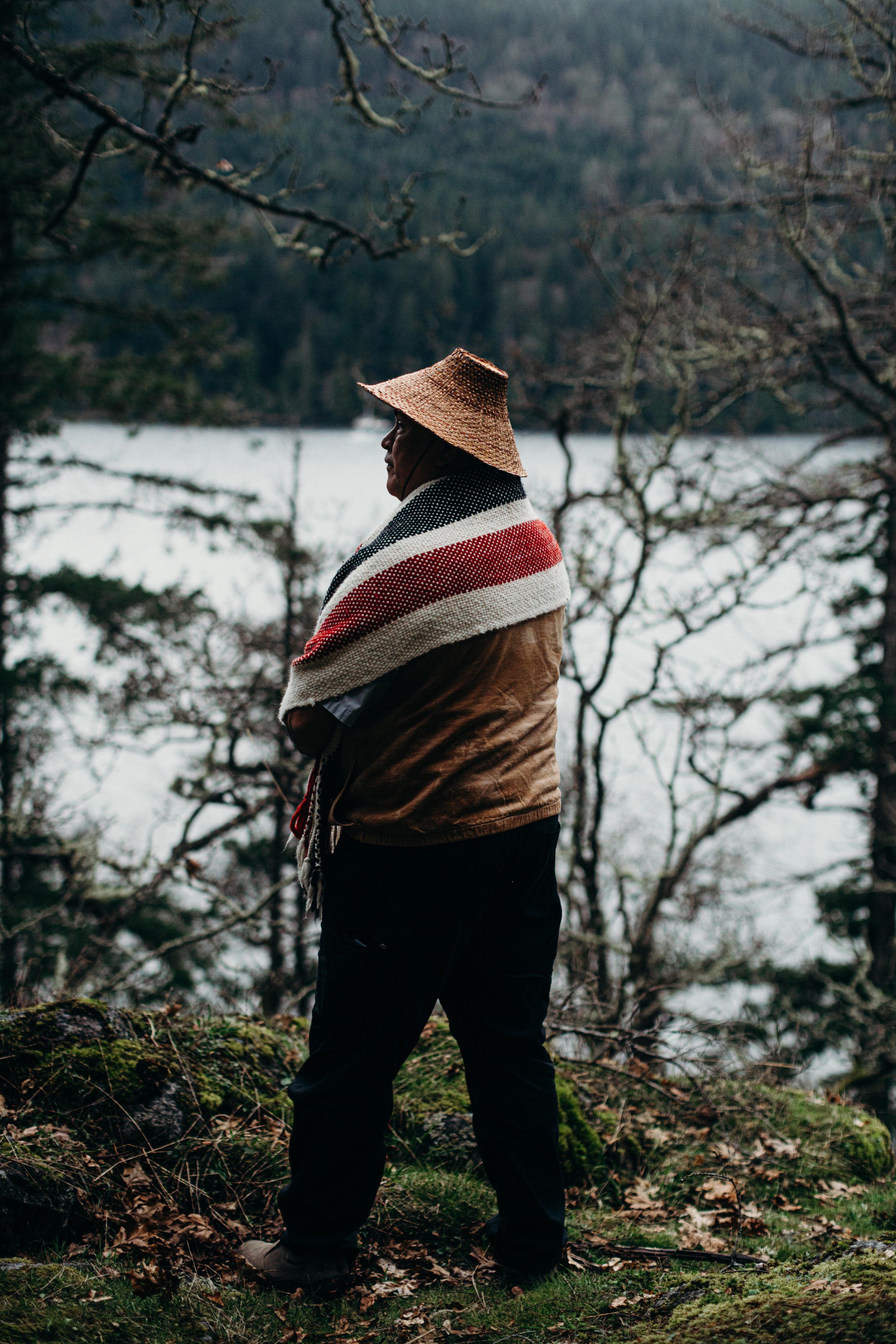 Man in straw hat and striped blanket stands on mossy ground overlooking a misty lake surrounded by bare trees
