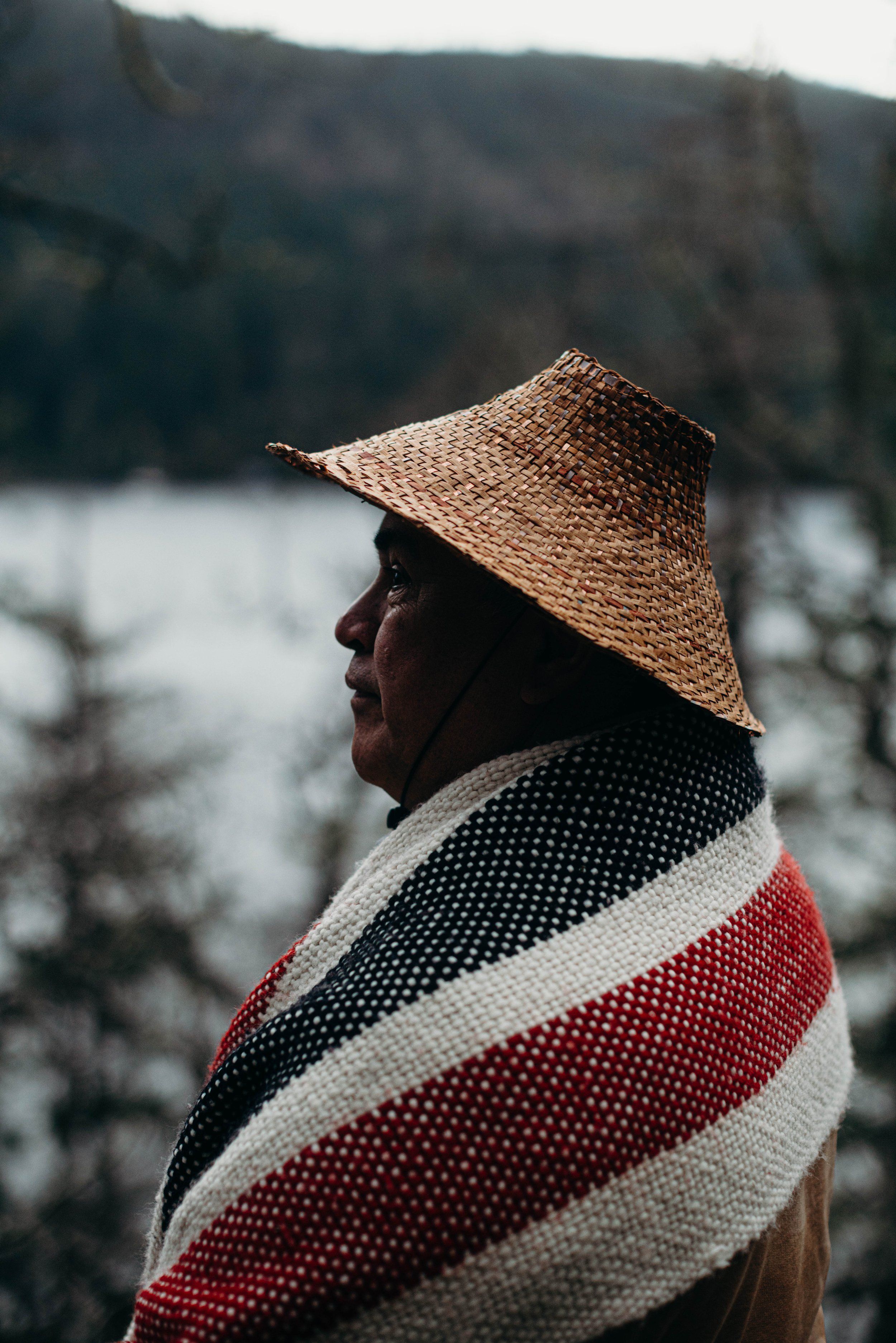 Man in straw hat and striped blanket gazes toward misty water and forested hills at golden hour