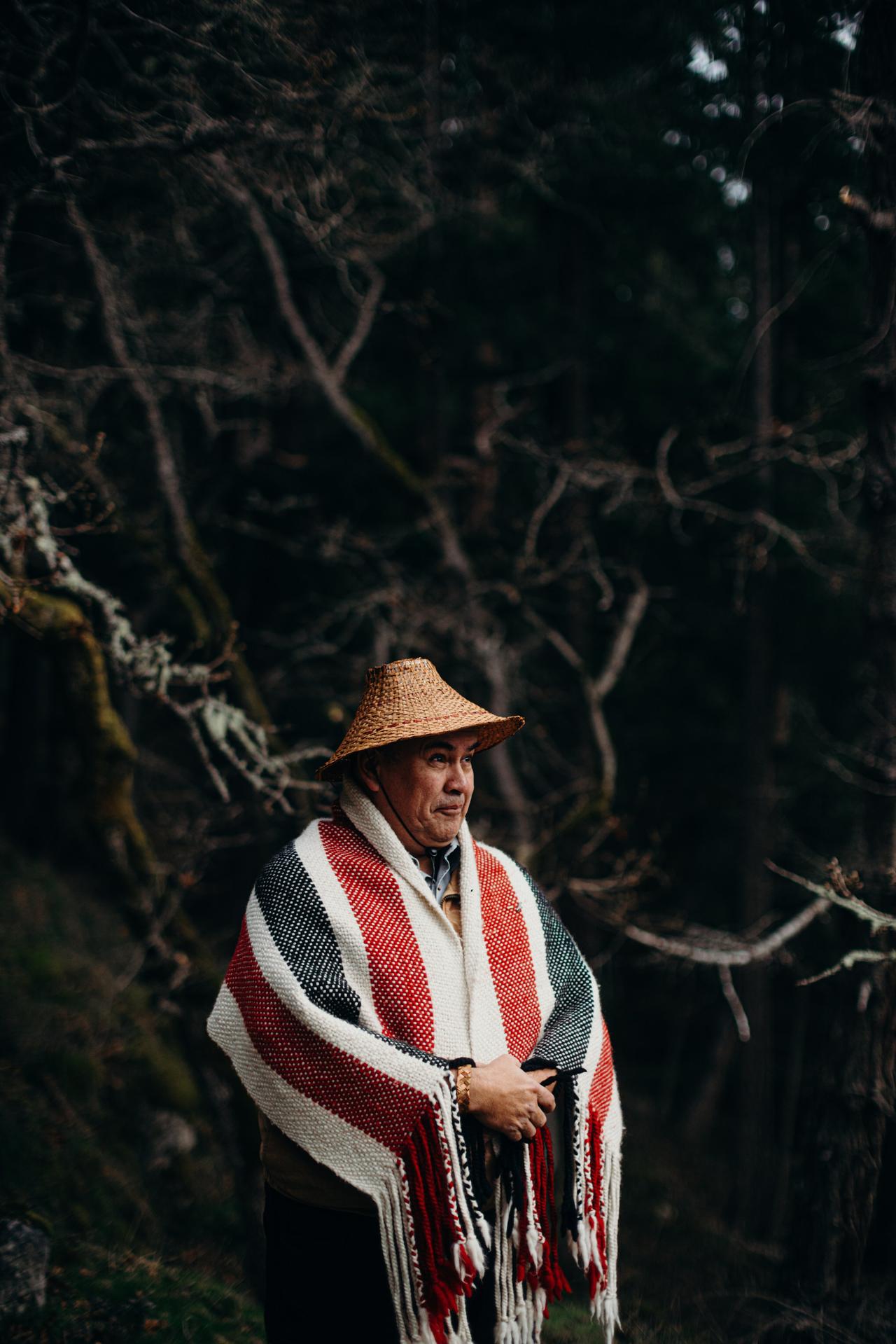 Man wearing woven hat and red-and-white striped blanket with fringe looks toward camera against dark forest background