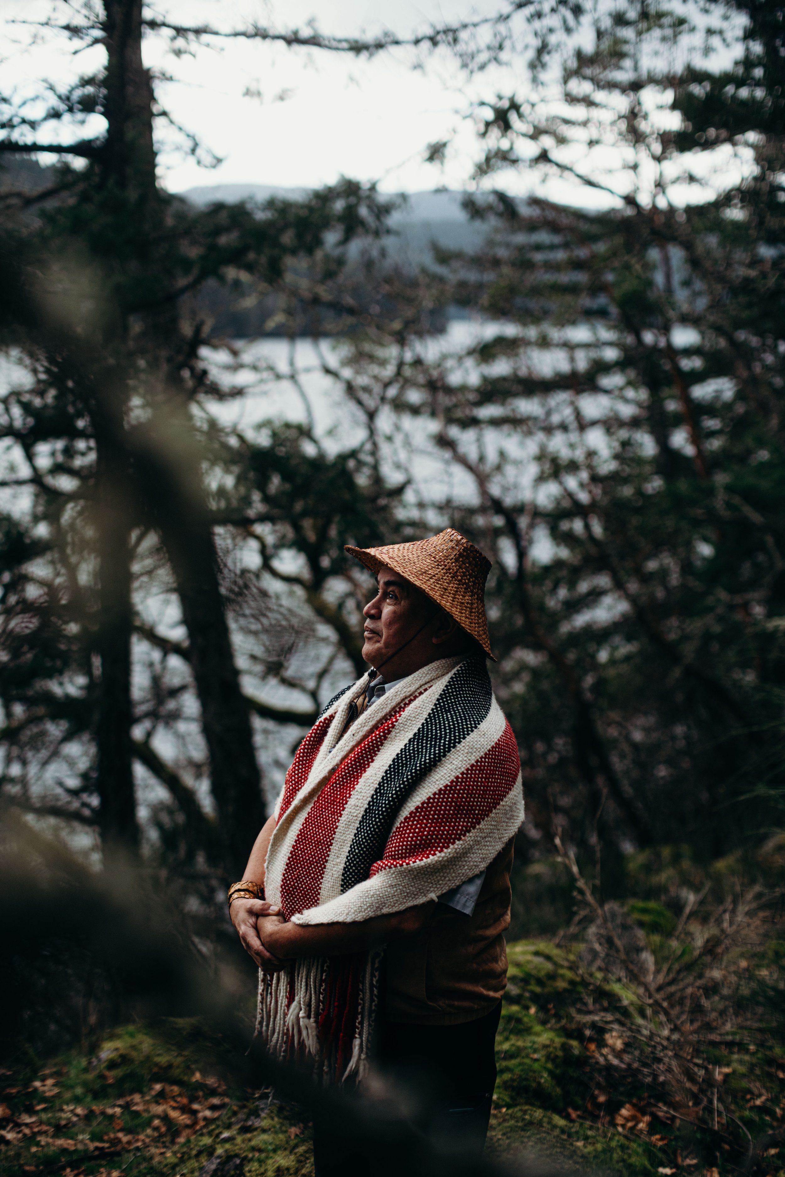 Man in straw hat and striped blanket gazes toward water framed by evergreen forest on misty day