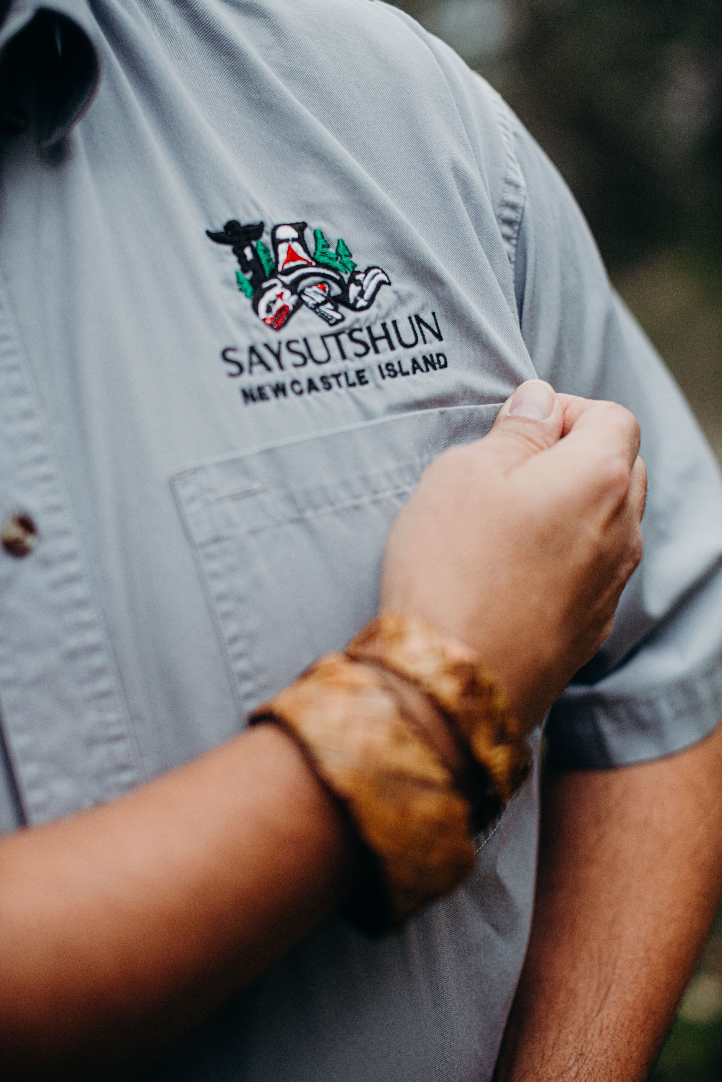 Man wearing Saysutshun Newcastle Island shirt holds fabric near chest, arm decorated with brown leather bracelet, soft natural light
