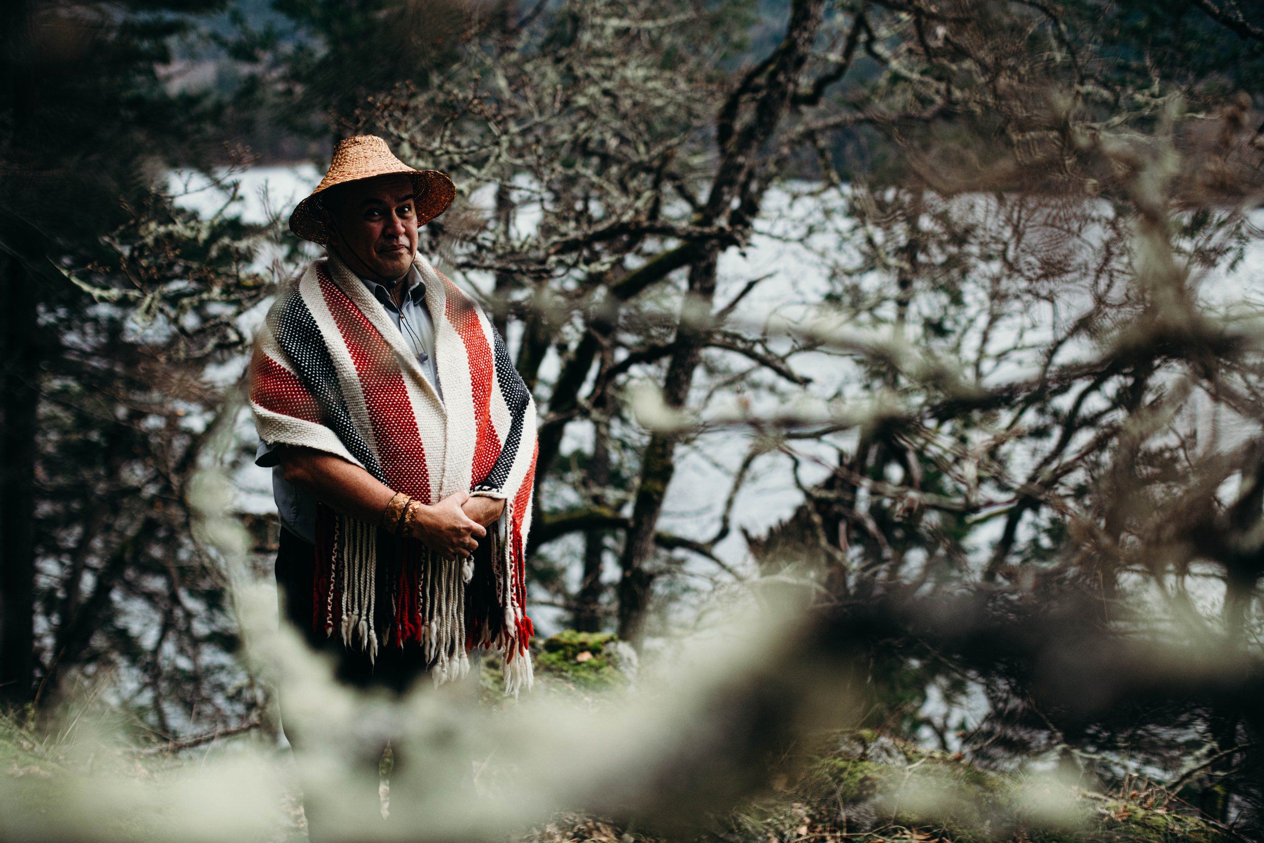 Man in wide-brimmed hat and red striped shawl stands among gnarled trees, gazing thoughtfully into distance