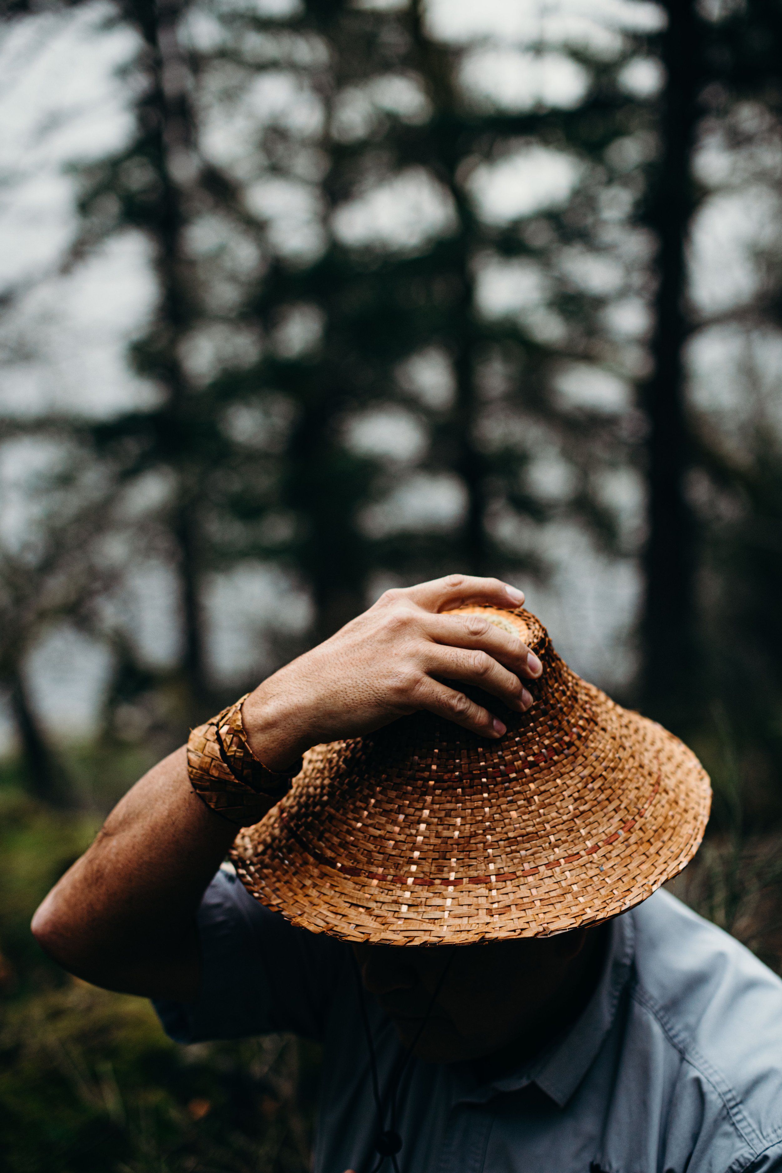 Person in blue shirt holds woven straw hat against face, hand resting on brim, with blurred forest trees behind in natural daylight