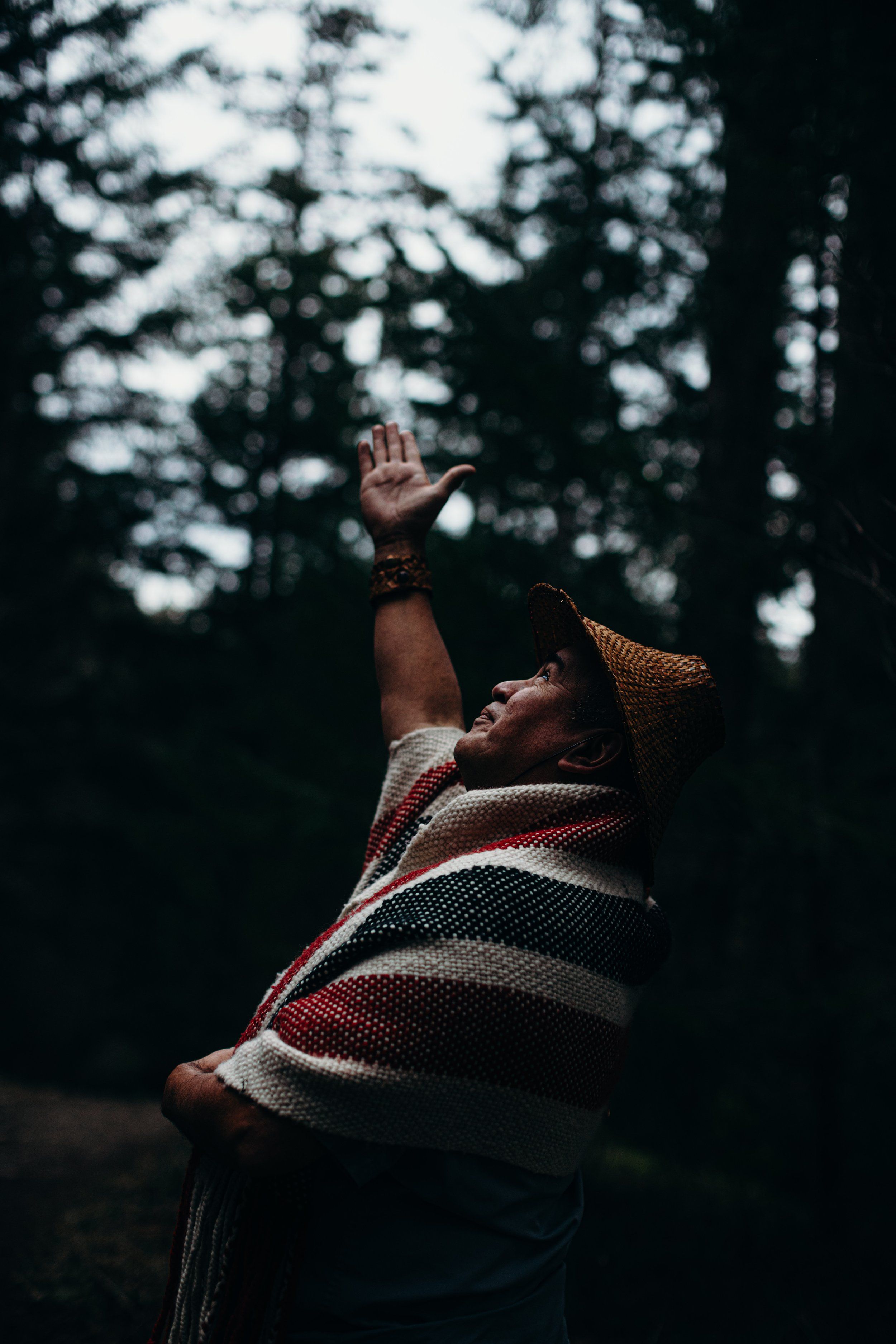 Man in striped blanket reaches skyward through dappled forest light, face tilted upward in contemplative gesture