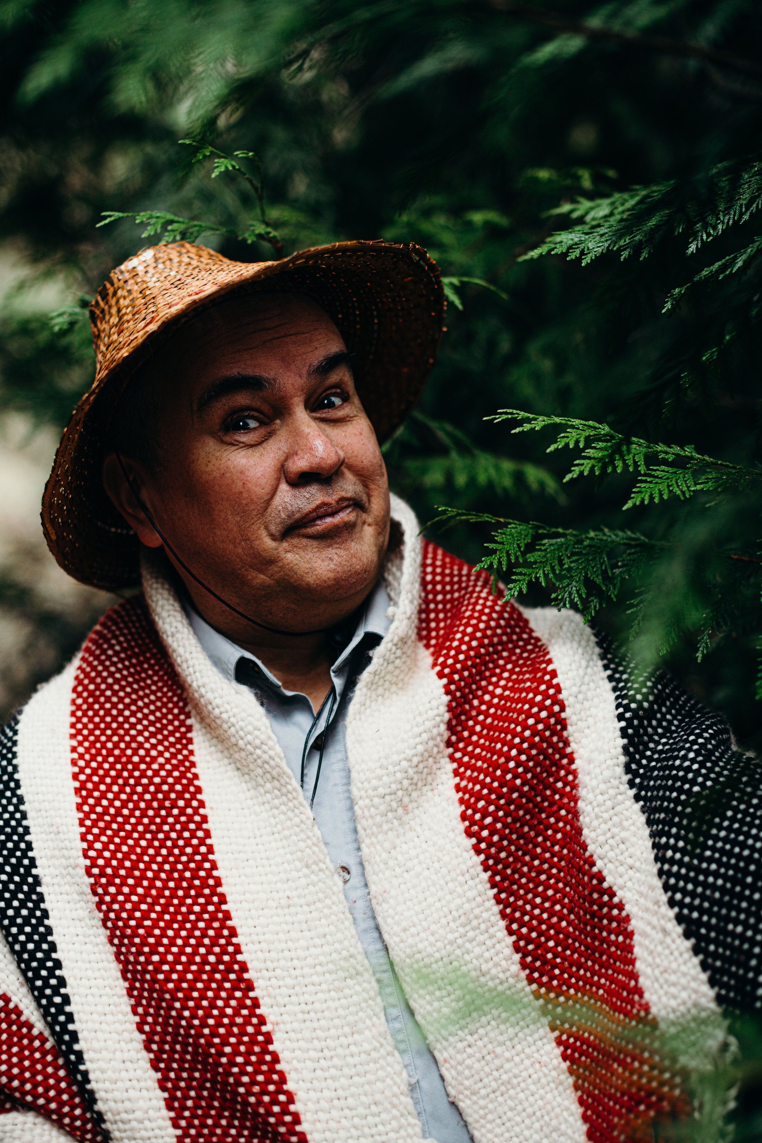 Man in woven hat and red-and-white striped blanket gazes upward among green foliage in natural light