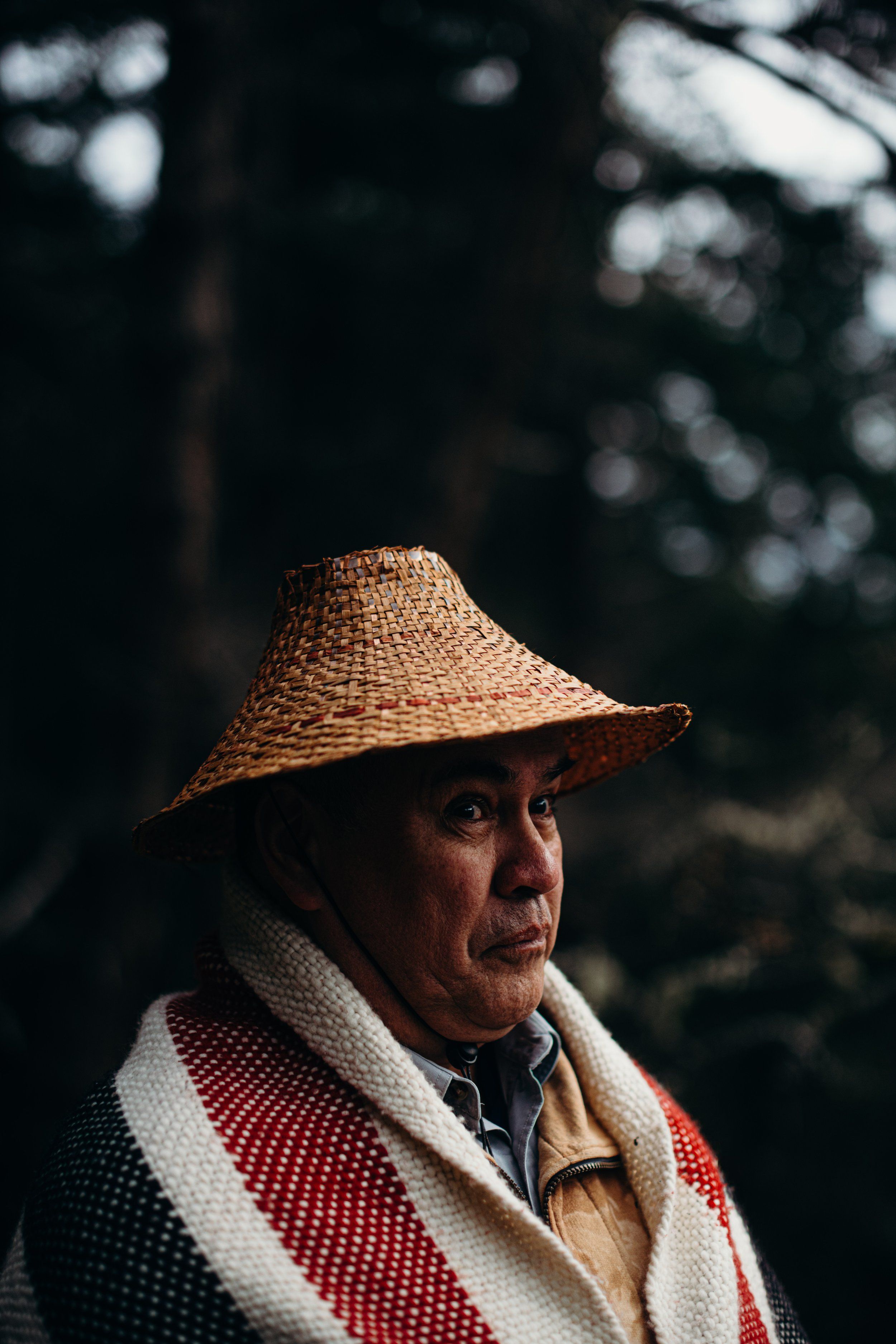 Indigenous man in woven hat and red-and-white blanket gazes thoughtfully into shadowed forest light