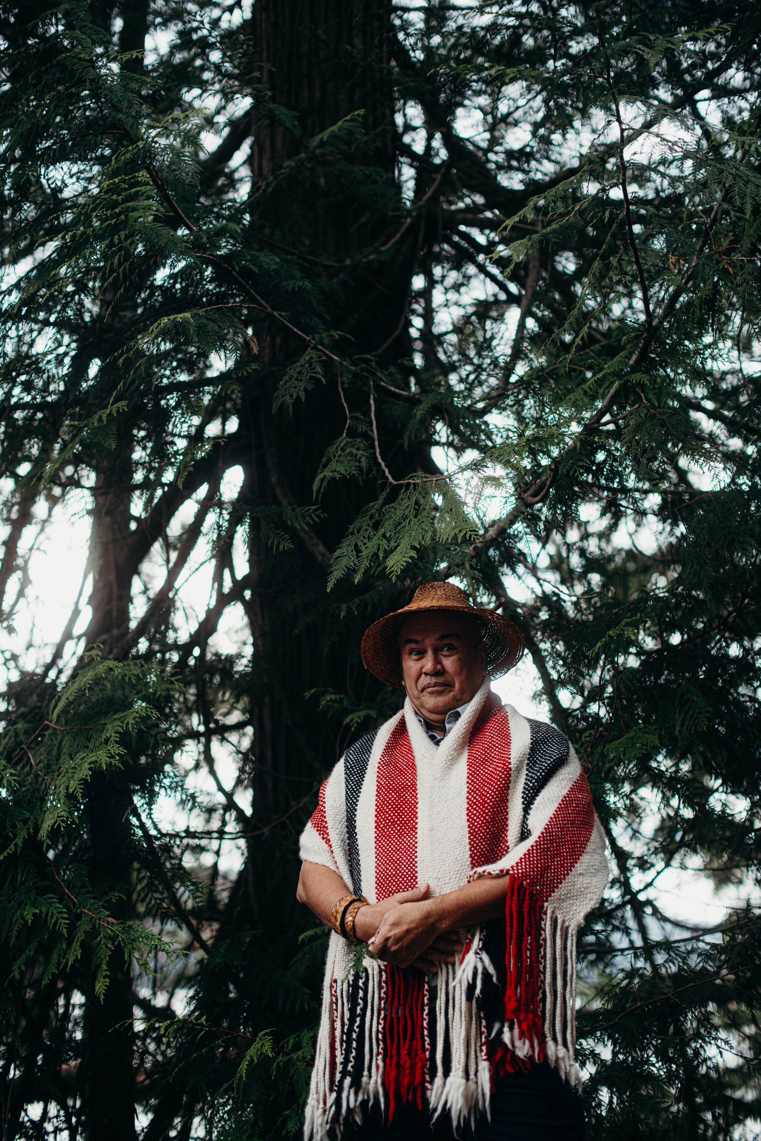 Man in brown hat and red-and-white striped blanket stands before towering evergreen trees in dappled forest light