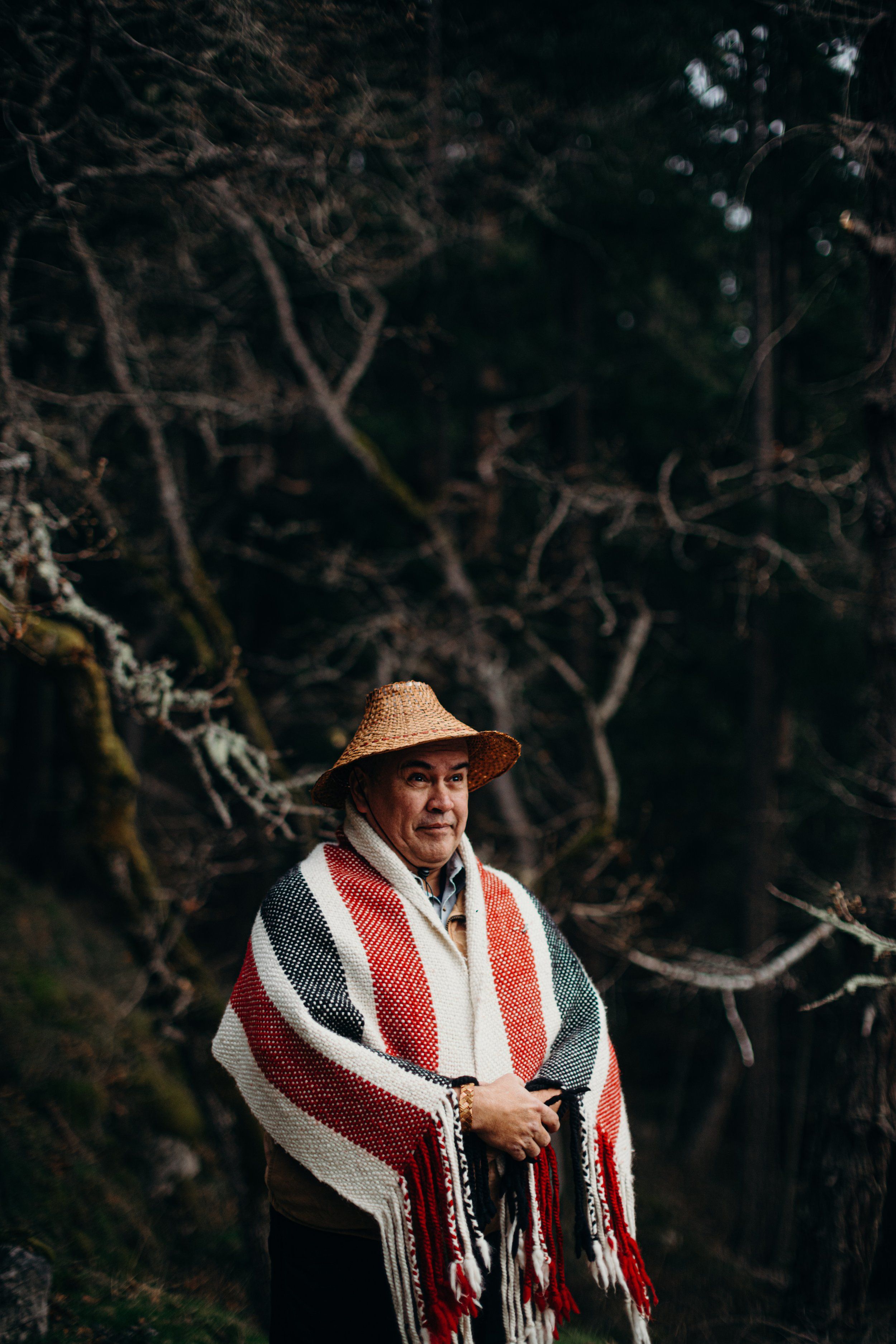 Indigenous man in woven blanket and straw hat stands contemplatively among bare winter branches in moody forest light