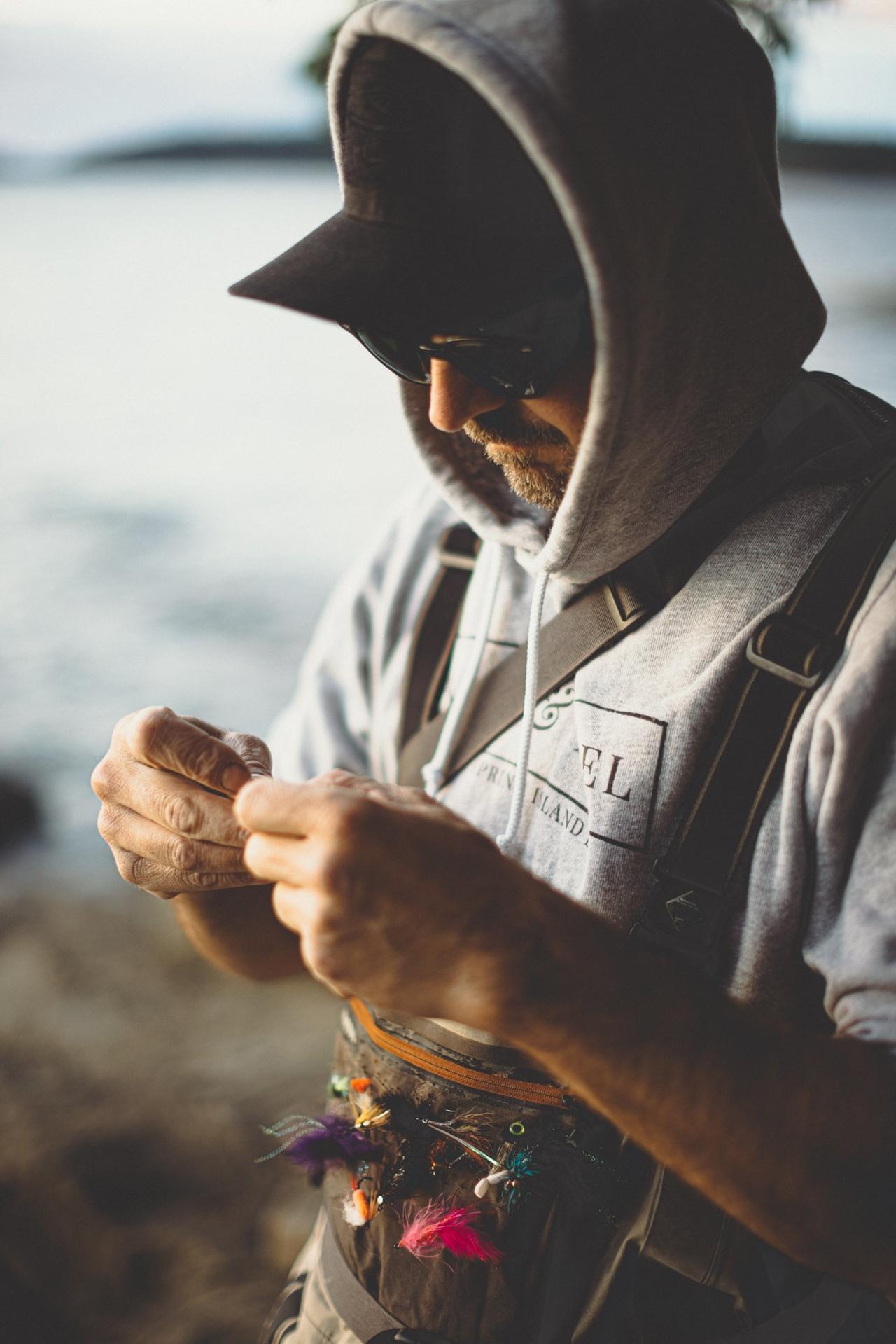 Bearded man in cowboy hat holds colorful fishing flies on a lanyard while wearing a hoodie