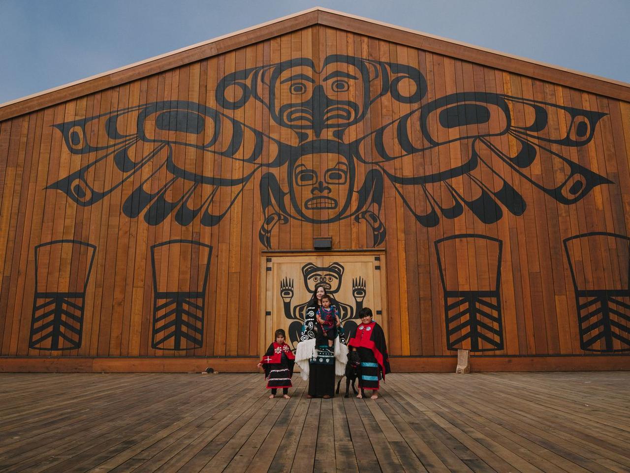 Four people stand before the Gwa'sala-'Nakwaxda'xw Nation big house, framed by formline art depicting faces and central
