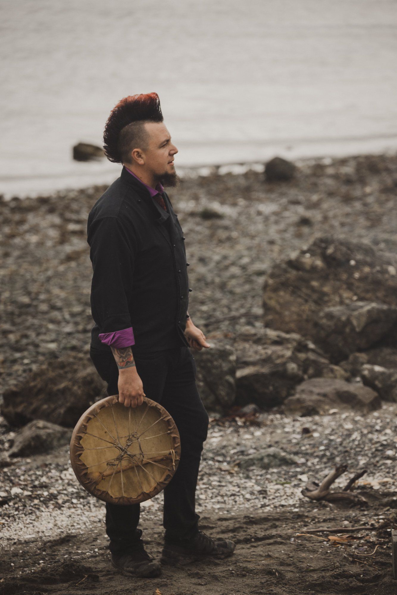 Man in black jacket holding wooden disc stands on rocky beach under overcast sky