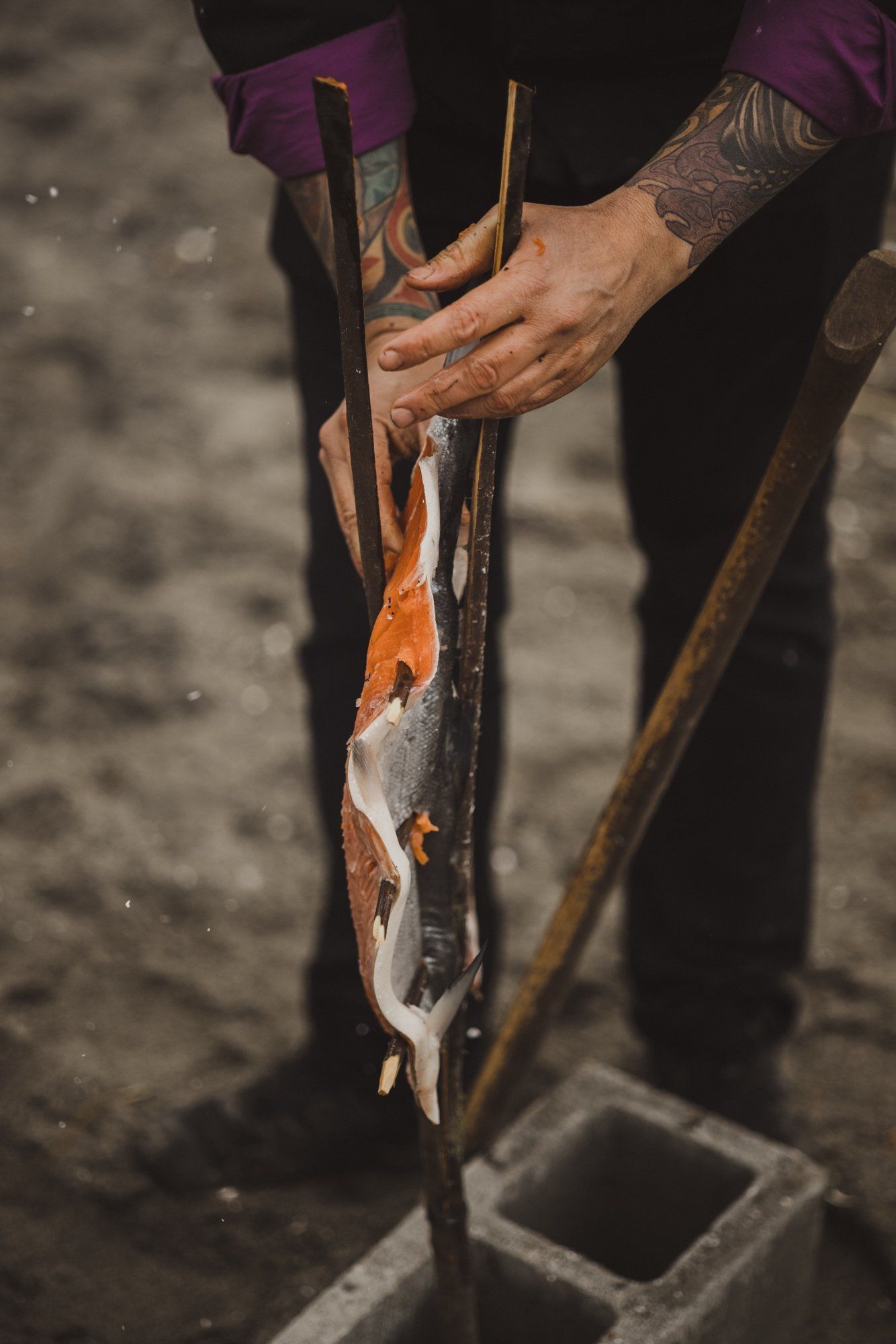 Tattooed hands holding a freshly caught fish over a wooden carving tool, coastal setting with natural light