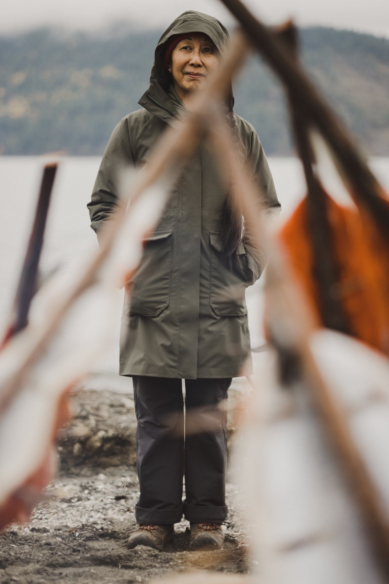 Young person in gray hoodie gazes upward by a beach, with blurred wooden logs in soft focus foreground