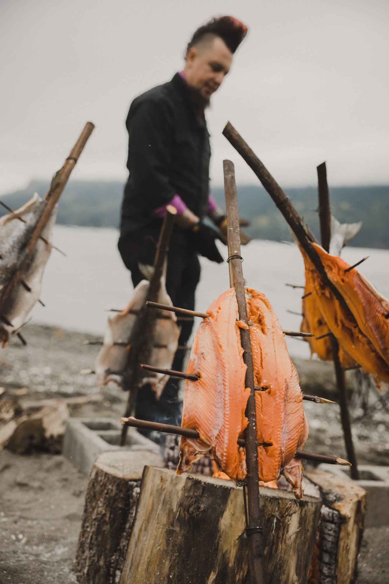 Man in black jacket stands beside smoked salmon on driftwood by misty water