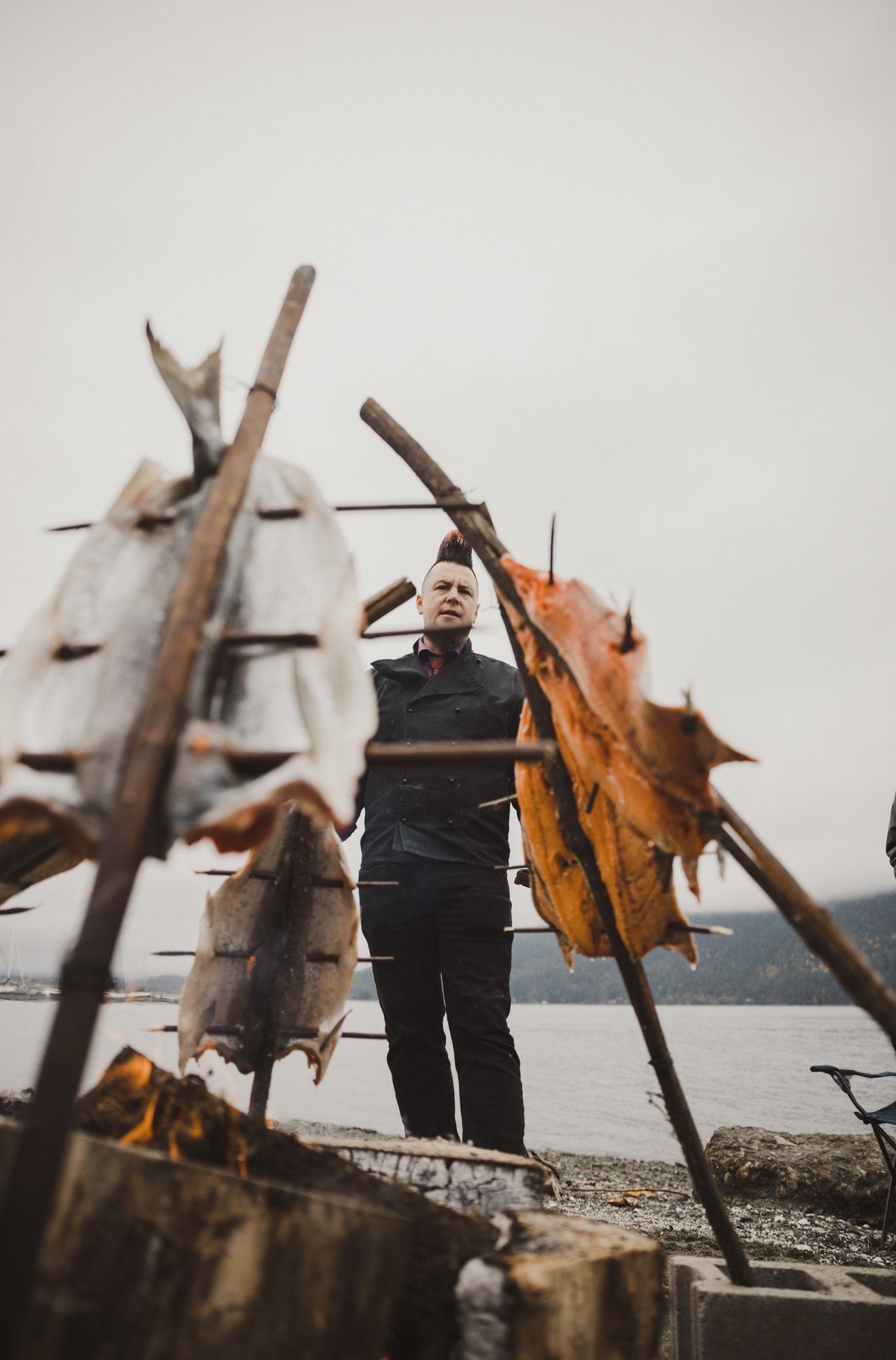 Man in black jacket reaching toward driftwood branches by misty water under overcast sky