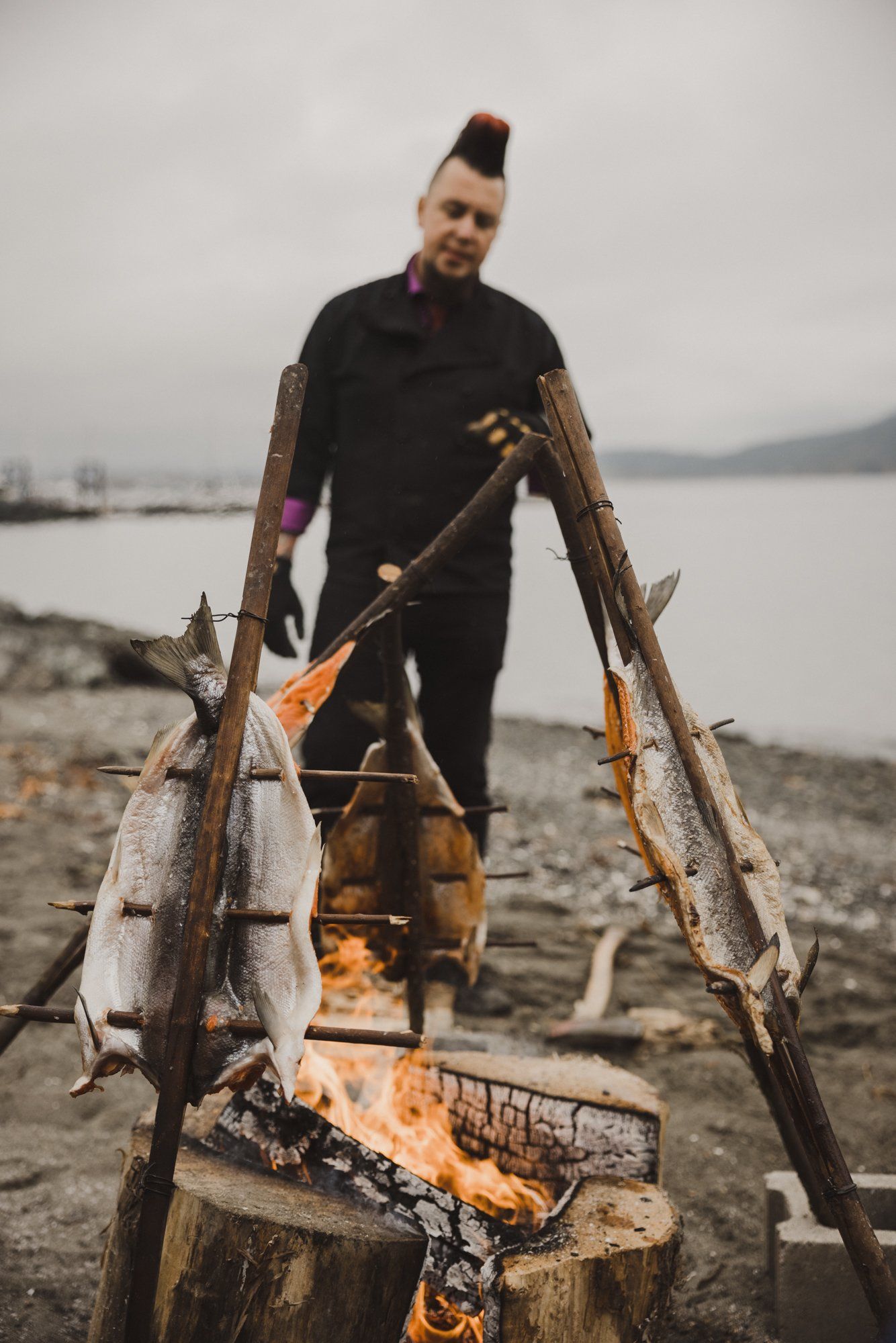 Man with mohawk holds freshly caught fish over driftwood fire on rocky beach with calm water beyond