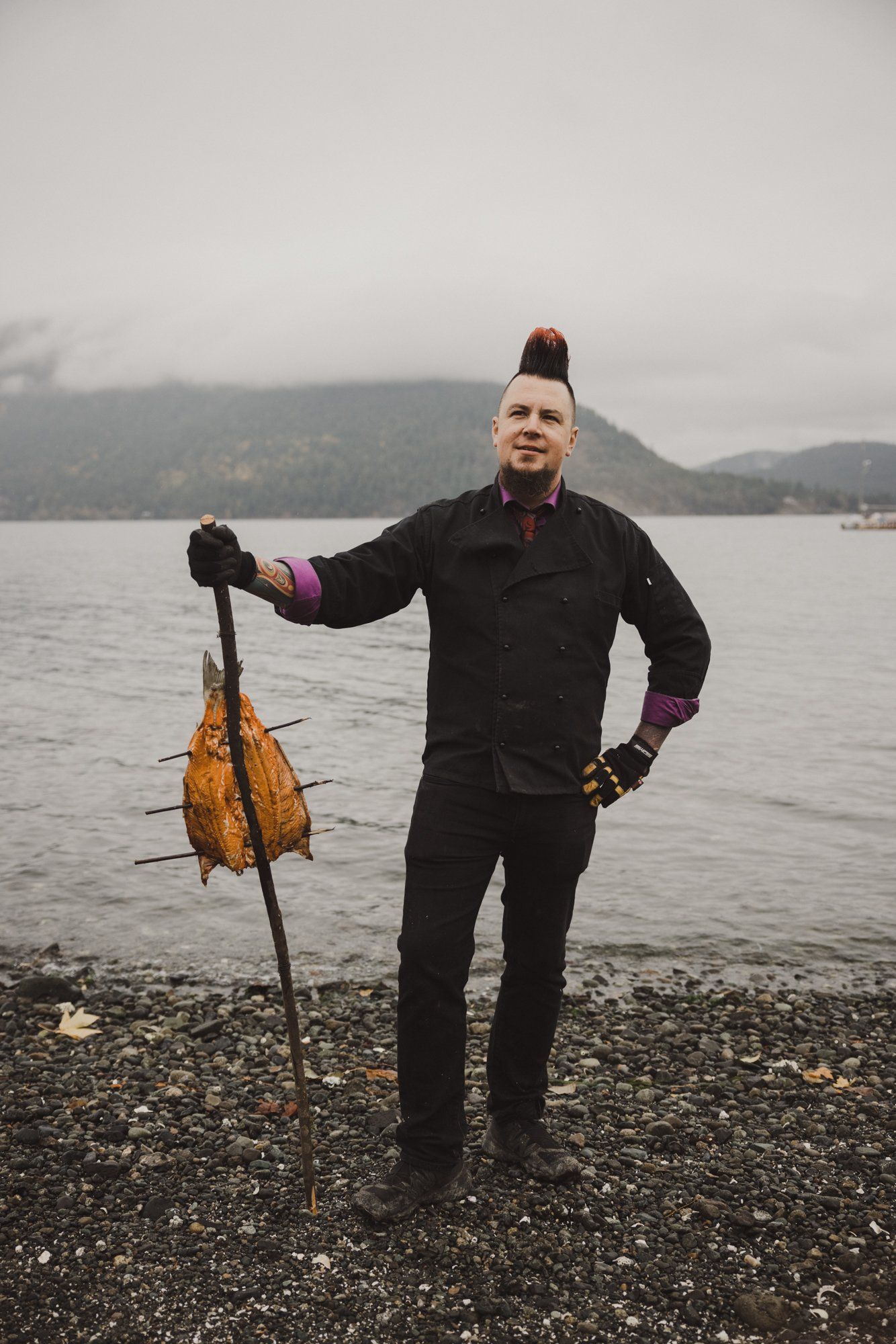 Man in black wetsuit holding harvested kelp on rocky beach with misty forested coastline behind