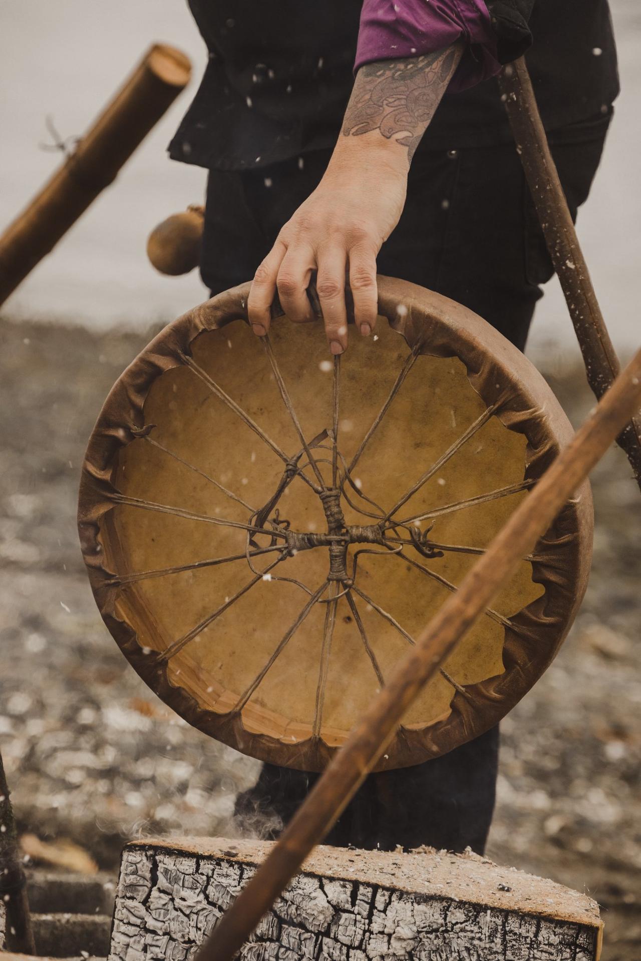 Hands holding a large traditional drum with rawhide head and wooden frame details, worn from use, against a dark background