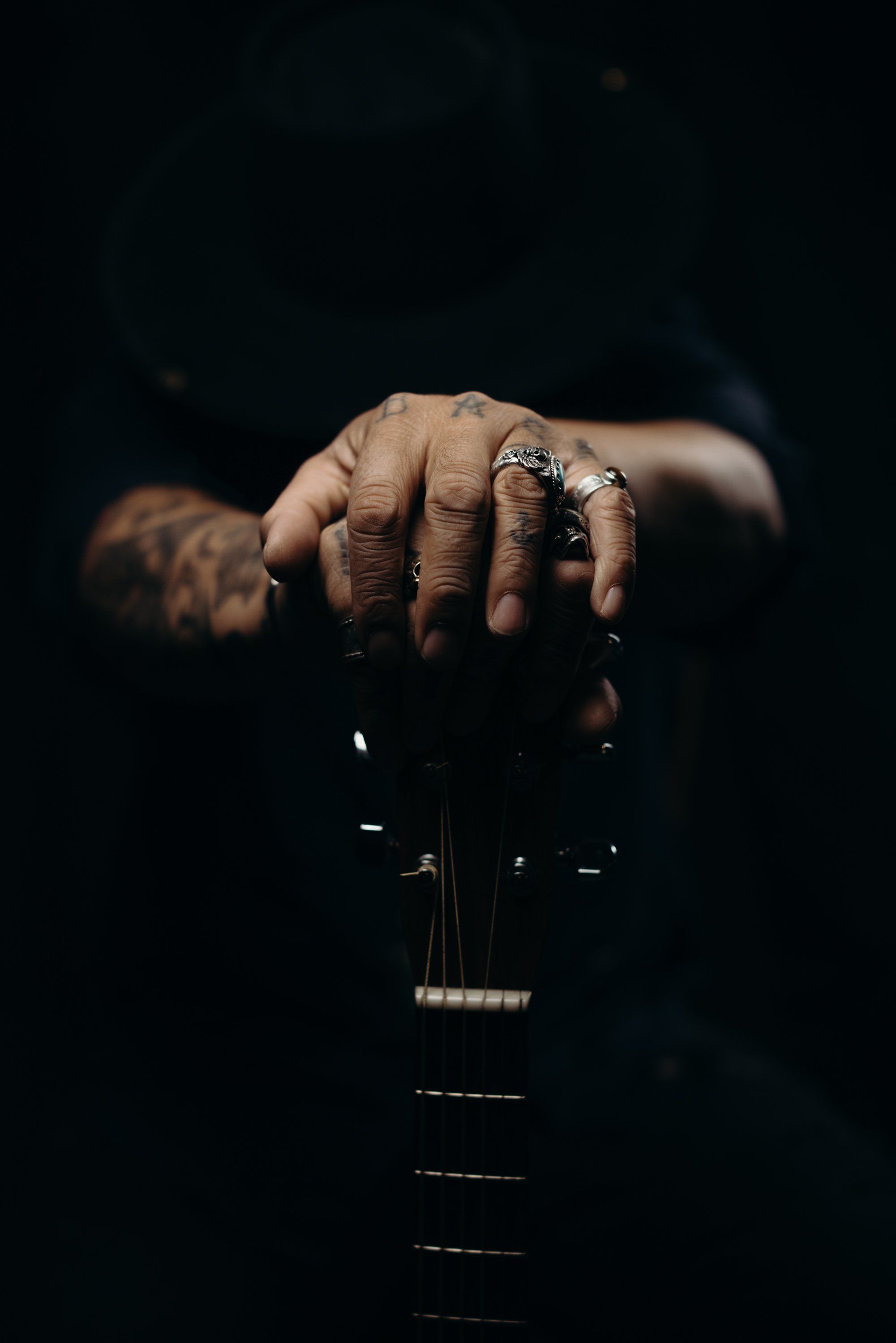 Tattooed hand with silver rings resting on guitar neck against dark background, intimate portrait lighting