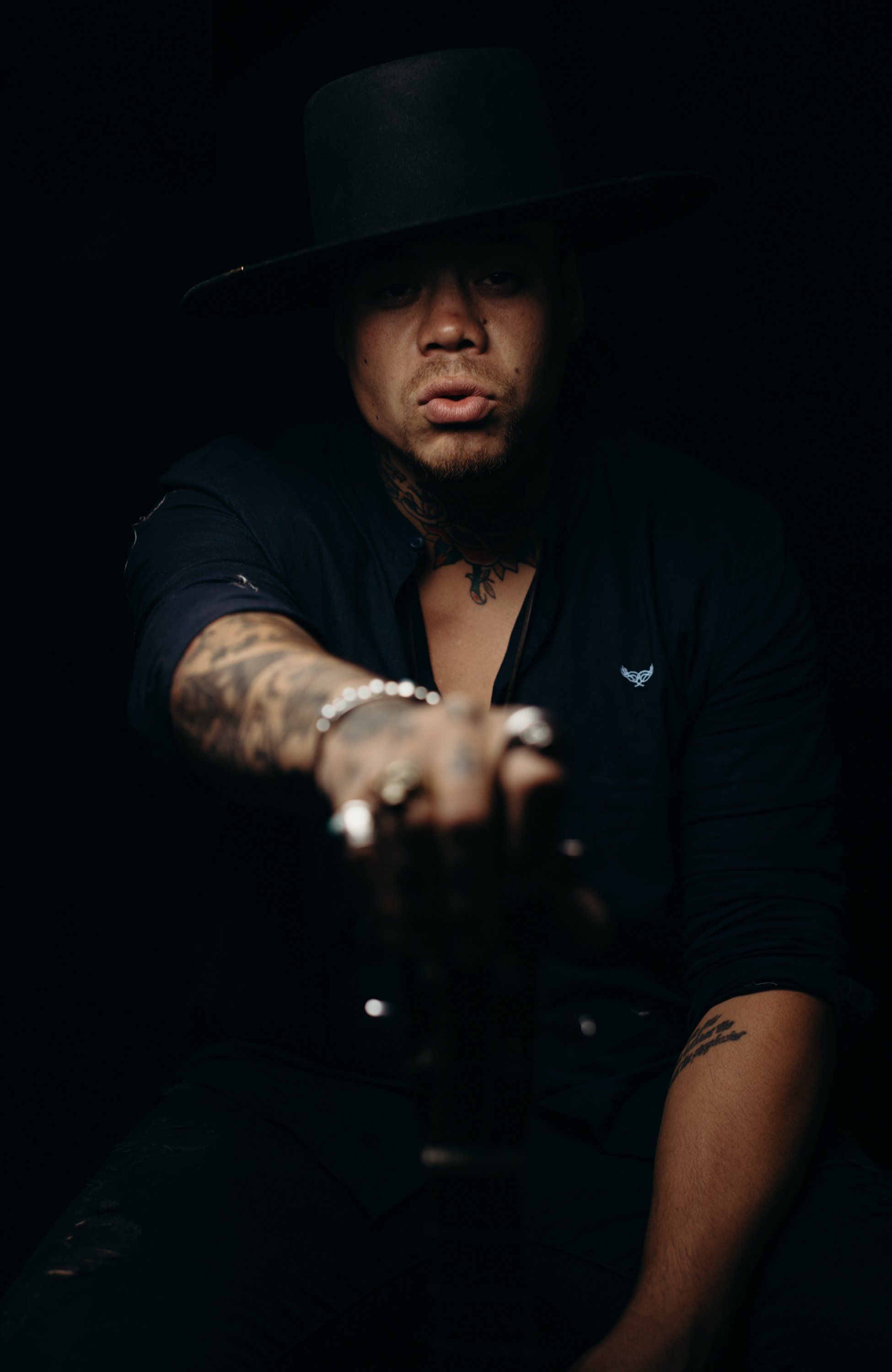 Man in black blazer and fedora pointing toward camera against dark background, dramatic side lighting