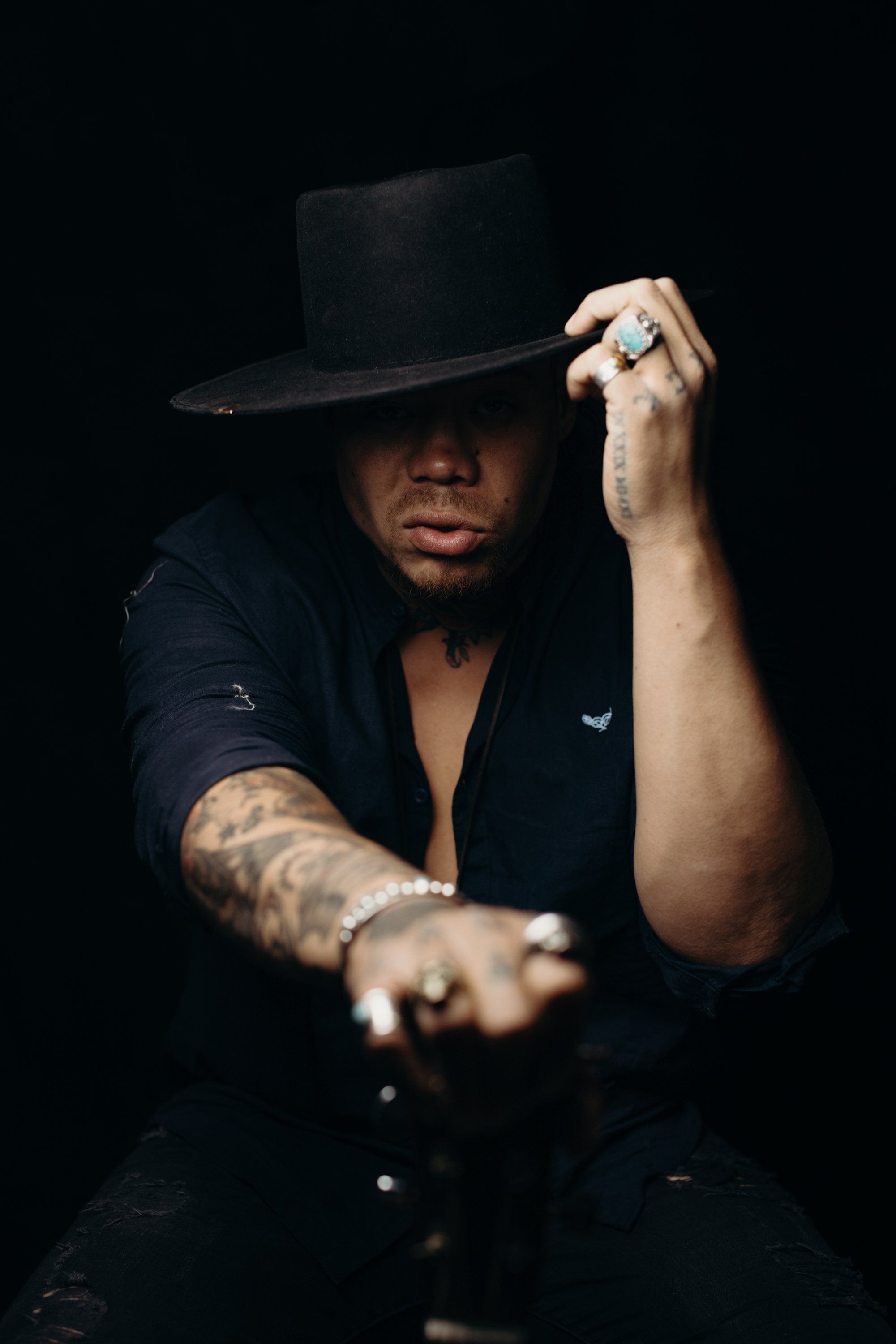 Person in black suit and fedora adjusting hat brim against dark background, arm extended toward camera with dramatic studio lighting