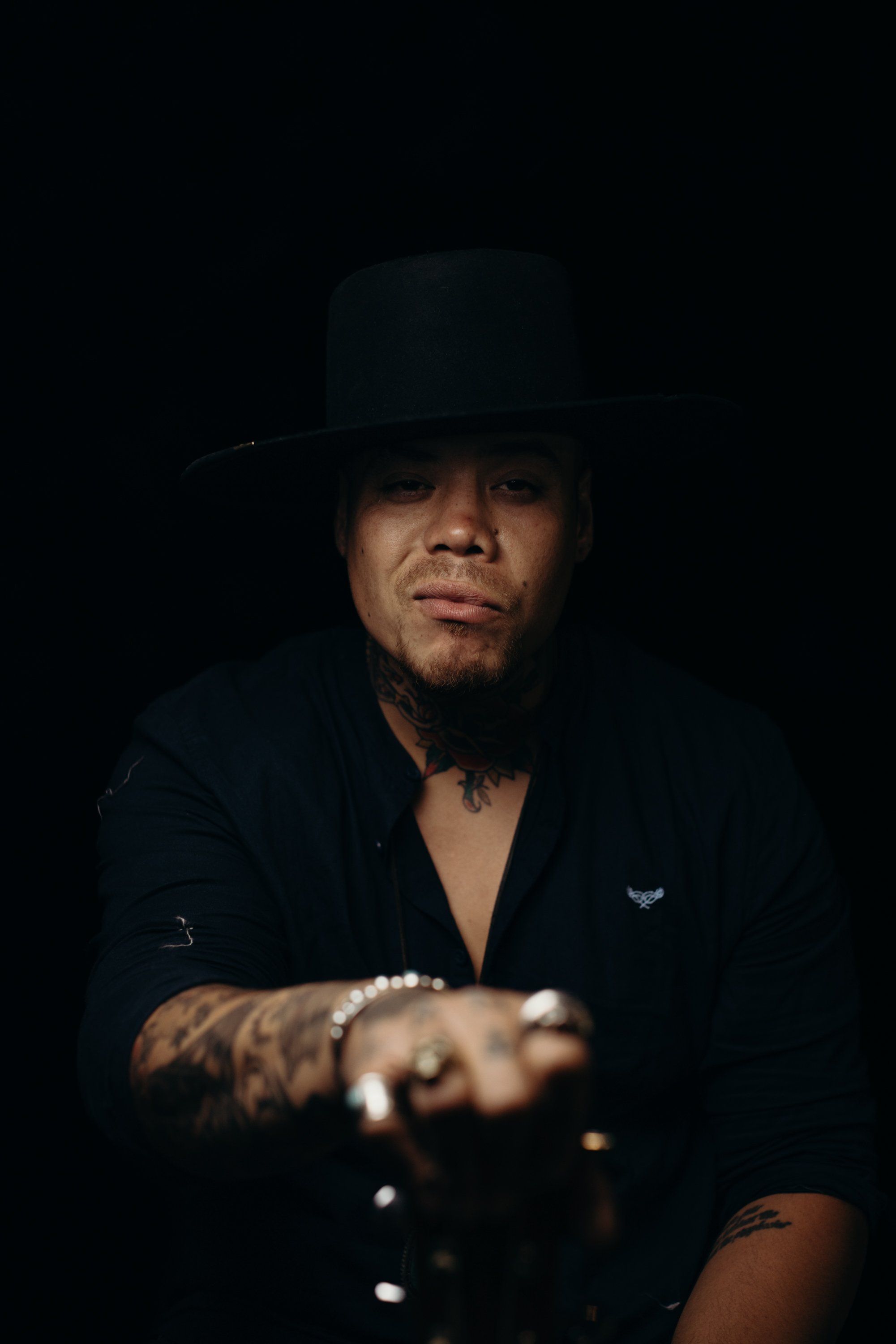 Person in black blazer and top hat against dark background, dramatic side lighting illuminating face and tattooed hand extended forward