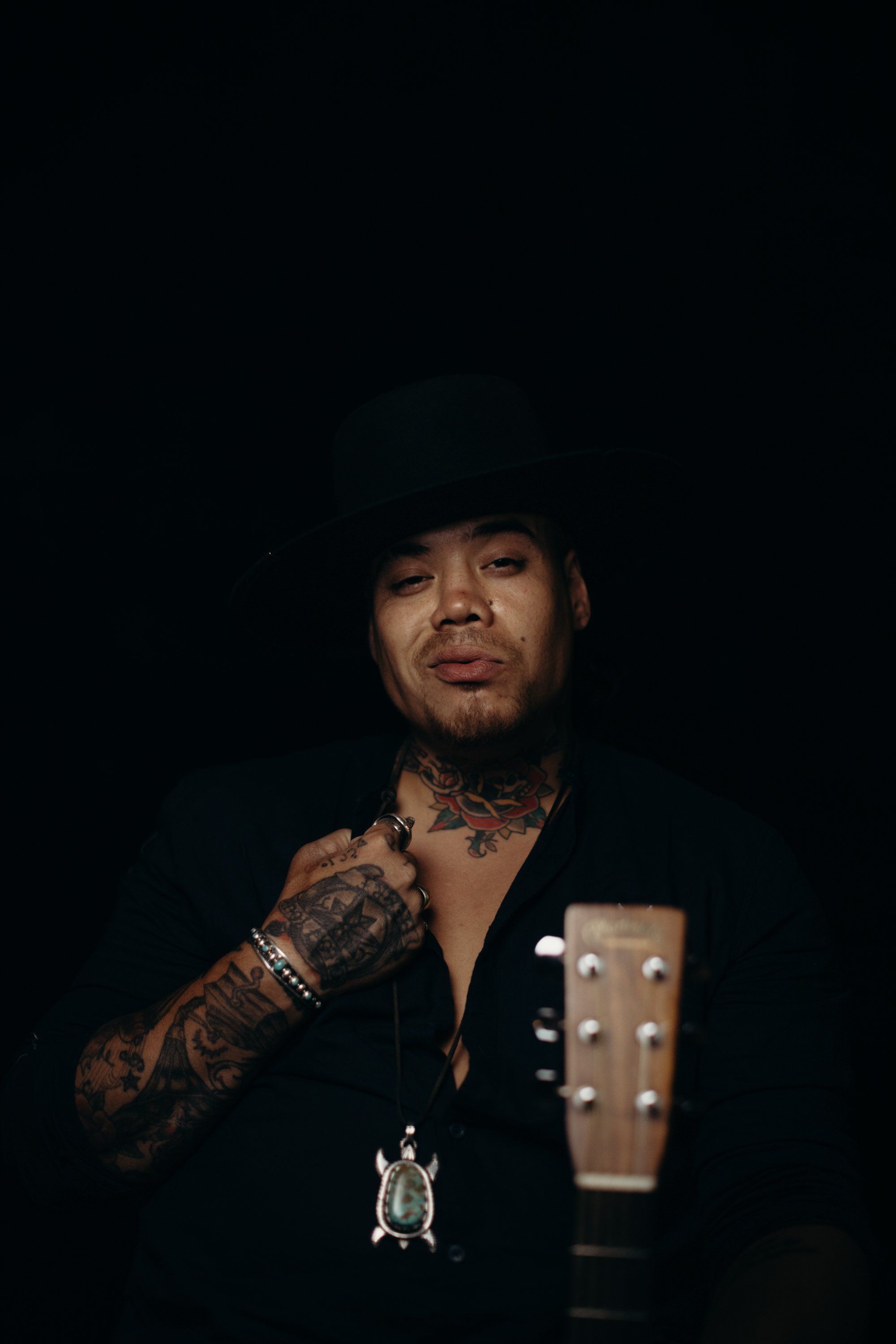 Tattooed musician in black jacket holds acoustic guitar against dark background, wearing gold chain and rings, direct gaze toward camera