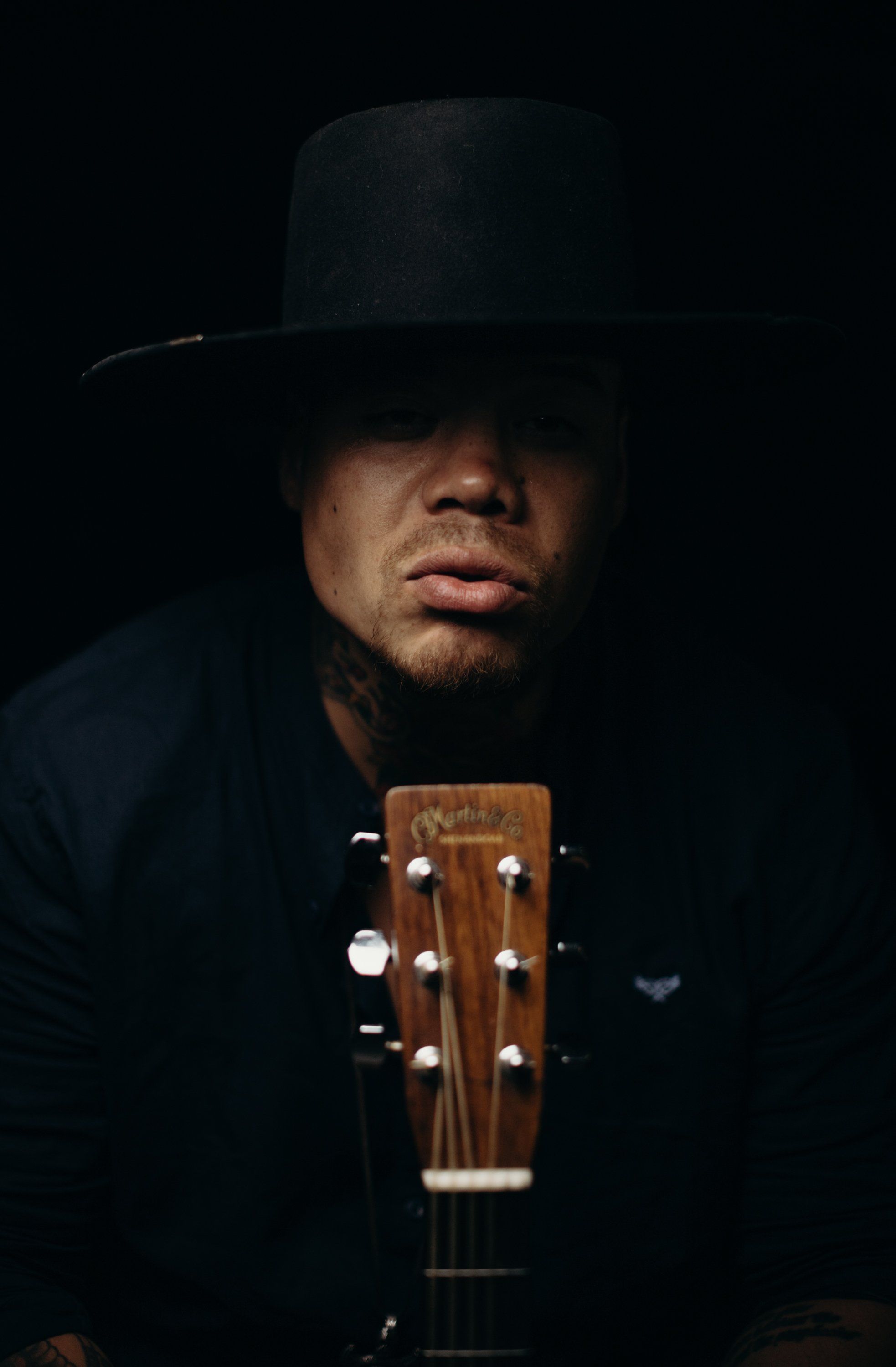 Man in black hat and jacket holds acoustic guitar against dark background, face partially shadowed in dramatic studio lighting