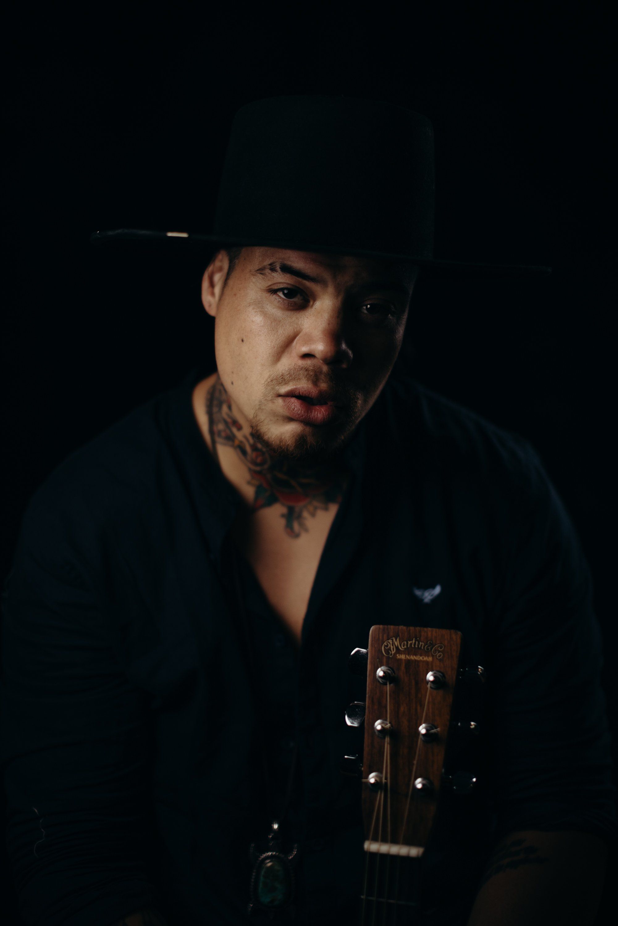 Man in black cowboy hat and jacket holding acoustic guitar against dark background, dramatic side lighting
