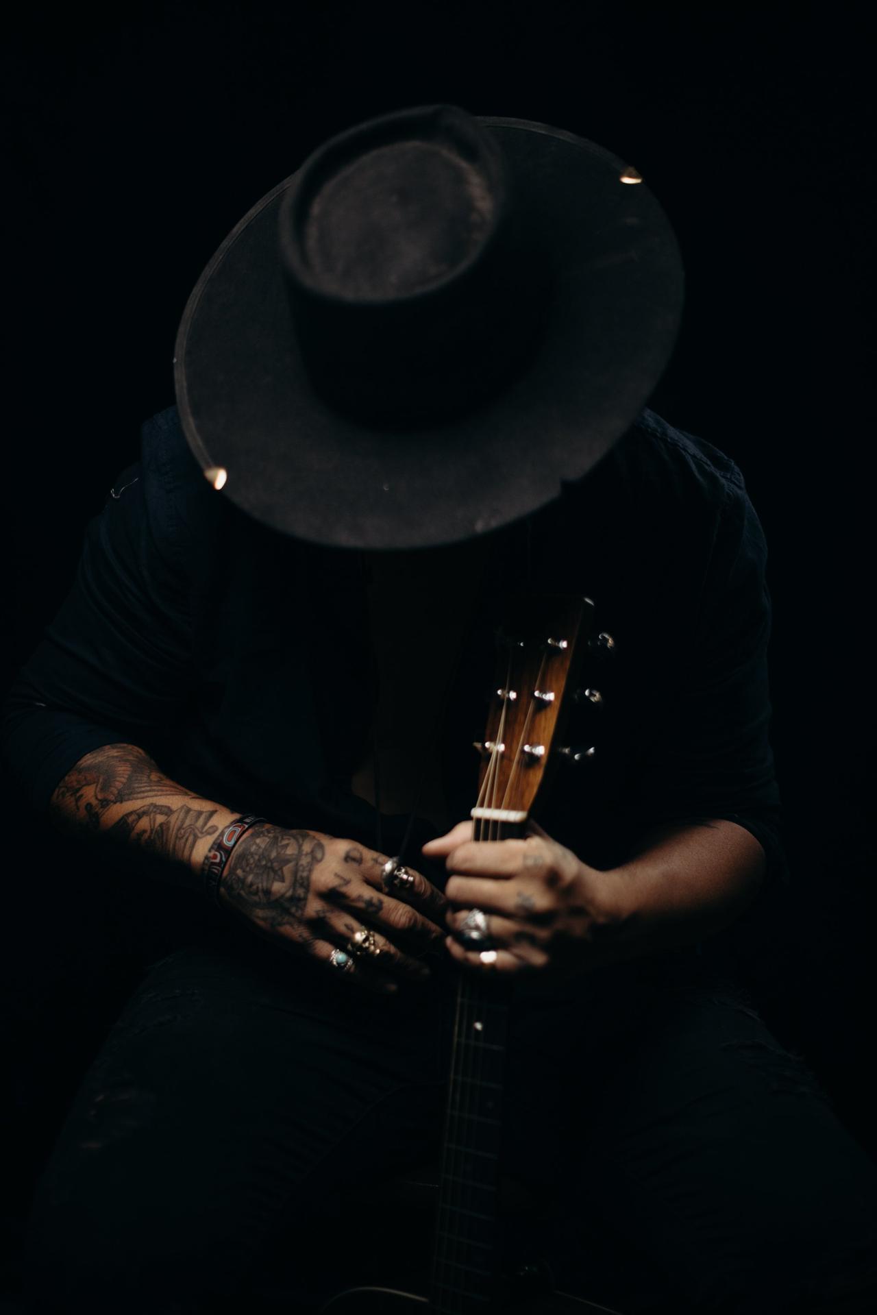 Tattooed hands with rings hold an acoustic guitar neck against a dark background, face obscured by shadow and guitar body